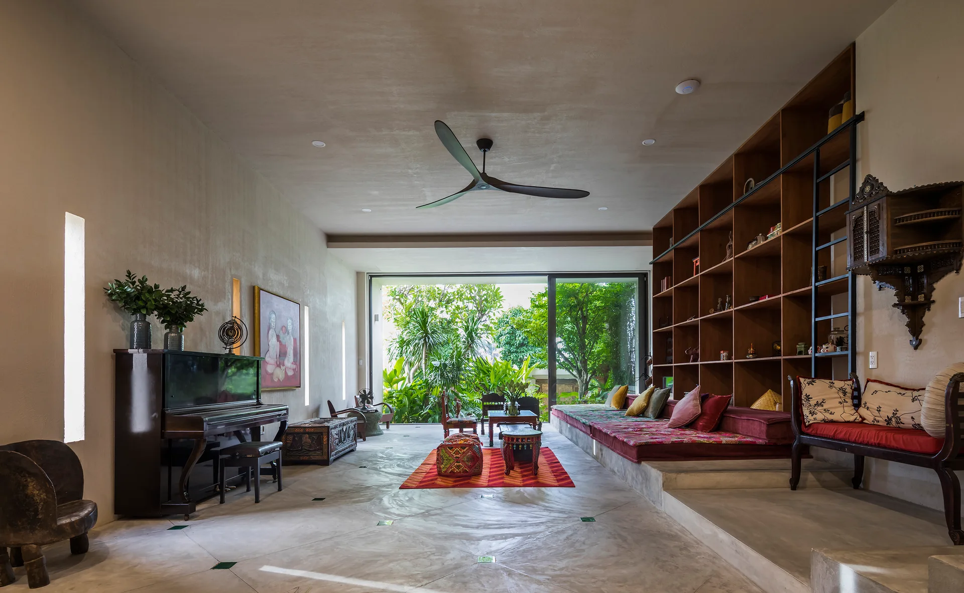 A spacious tropical living room features a concrete floor, built-in wooden bookcase with integrated daybed, and a glass door opening to a lush garden.