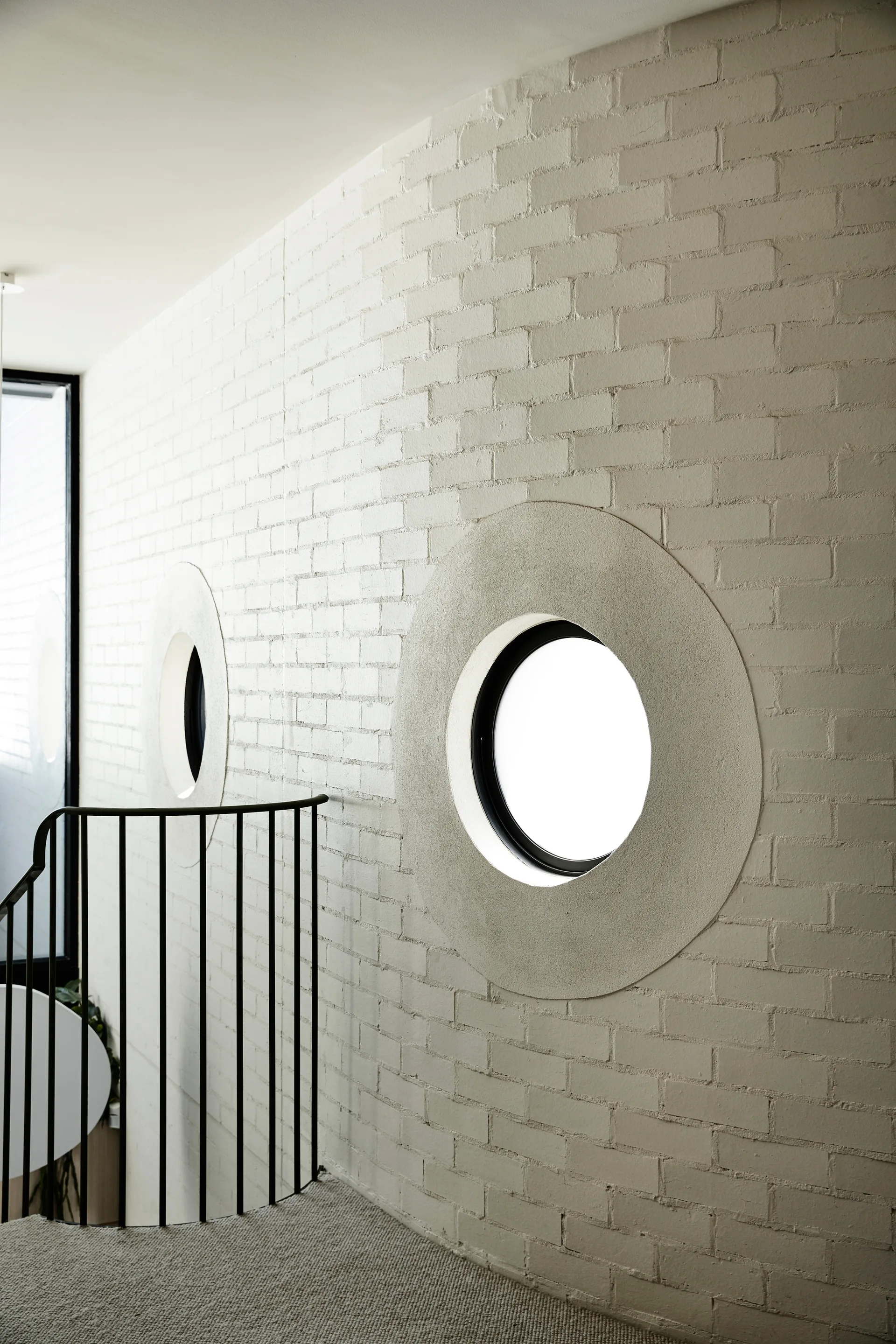 A white painted brick stairwell wall features two circular windows with smooth round borders next to a curved black metal railing.