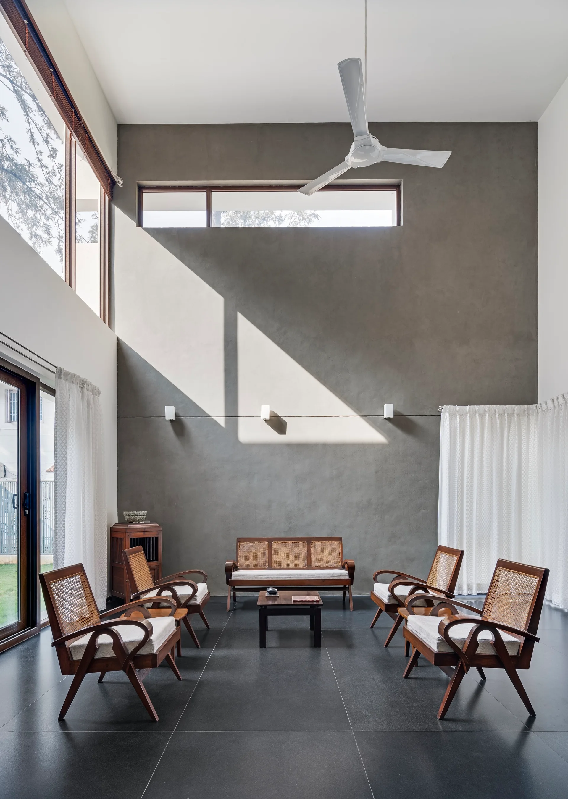 A spacious double-height living room featuring a tall grey concrete wall, symmetrical rattan and wood armchairs, a white ceiling fan, and dark tile floors.