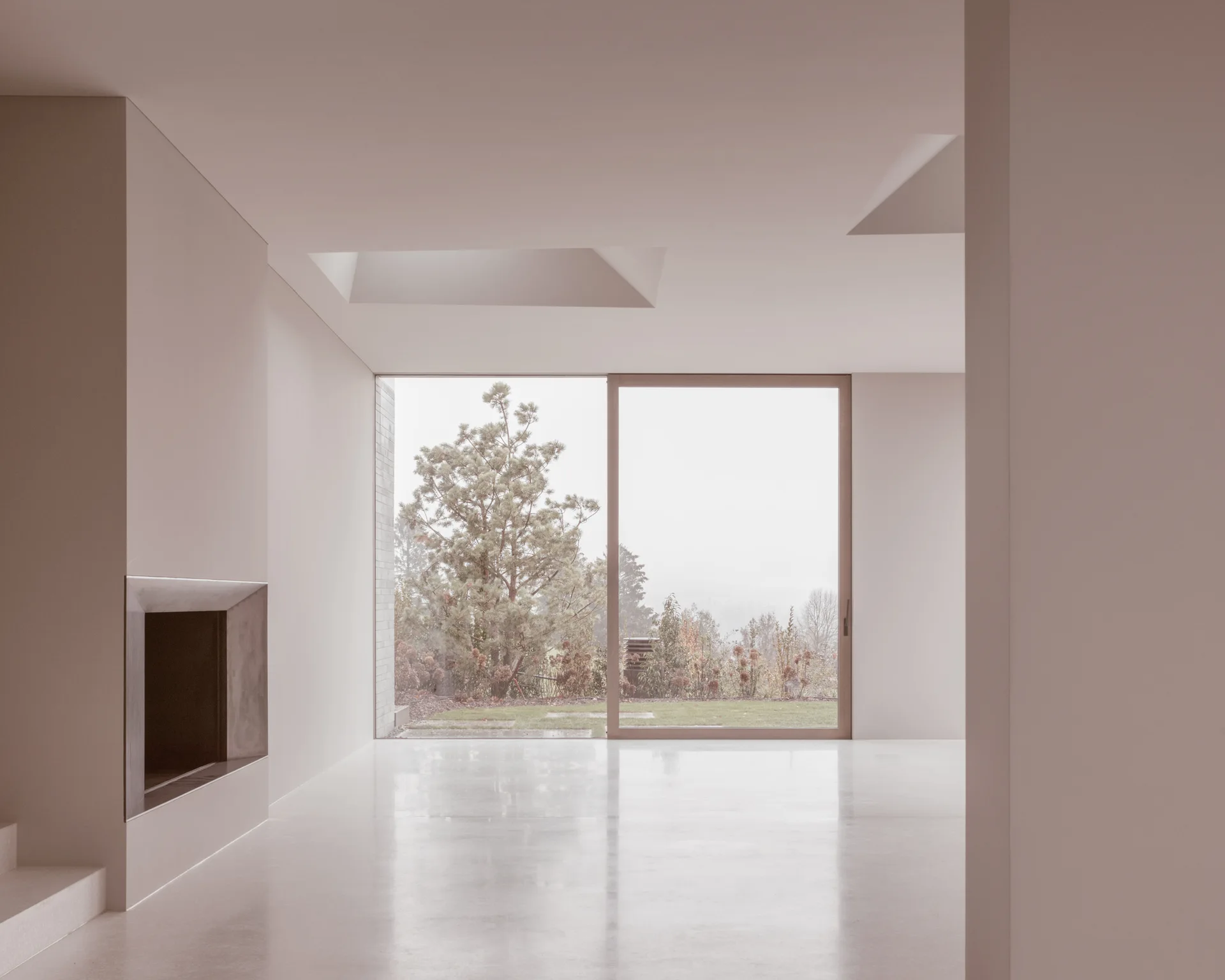 An empty minimalist room with warm beige walls, polished floors, a built-in fireplace, and a large sliding glass door opening to a garden.