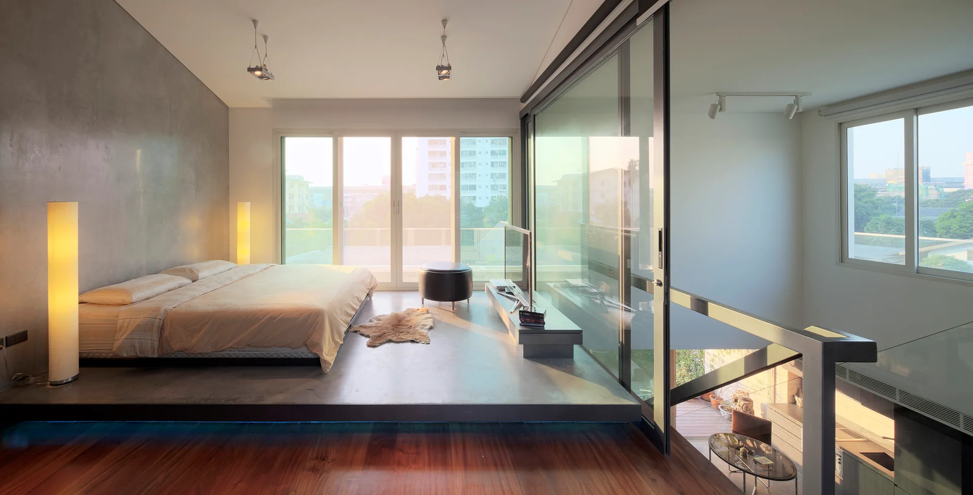 A minimalist bedroom features a low bed on a raised concrete platform next to sliding glass doors and a glass railing overlooking a lower level.