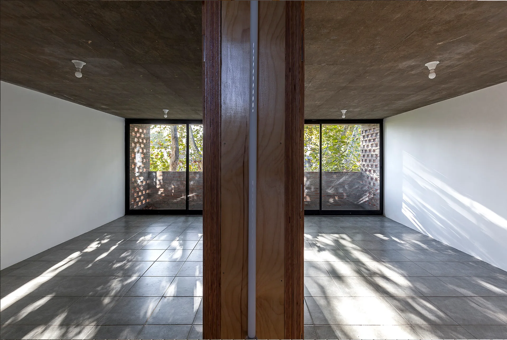 An empty minimalist room featuring a central wood partition, raw concrete ceiling, gray tiled floor, and large sliding glass windows.