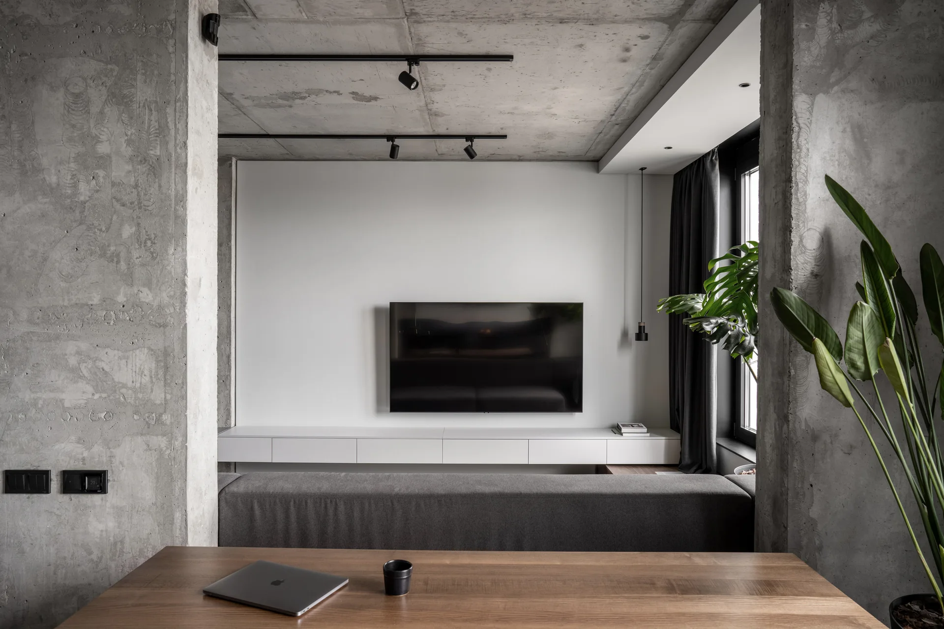 A modern living room viewed from a wooden desk, featuring raw concrete walls, a grey sofa, a wall-mounted TV, and a white floating console.