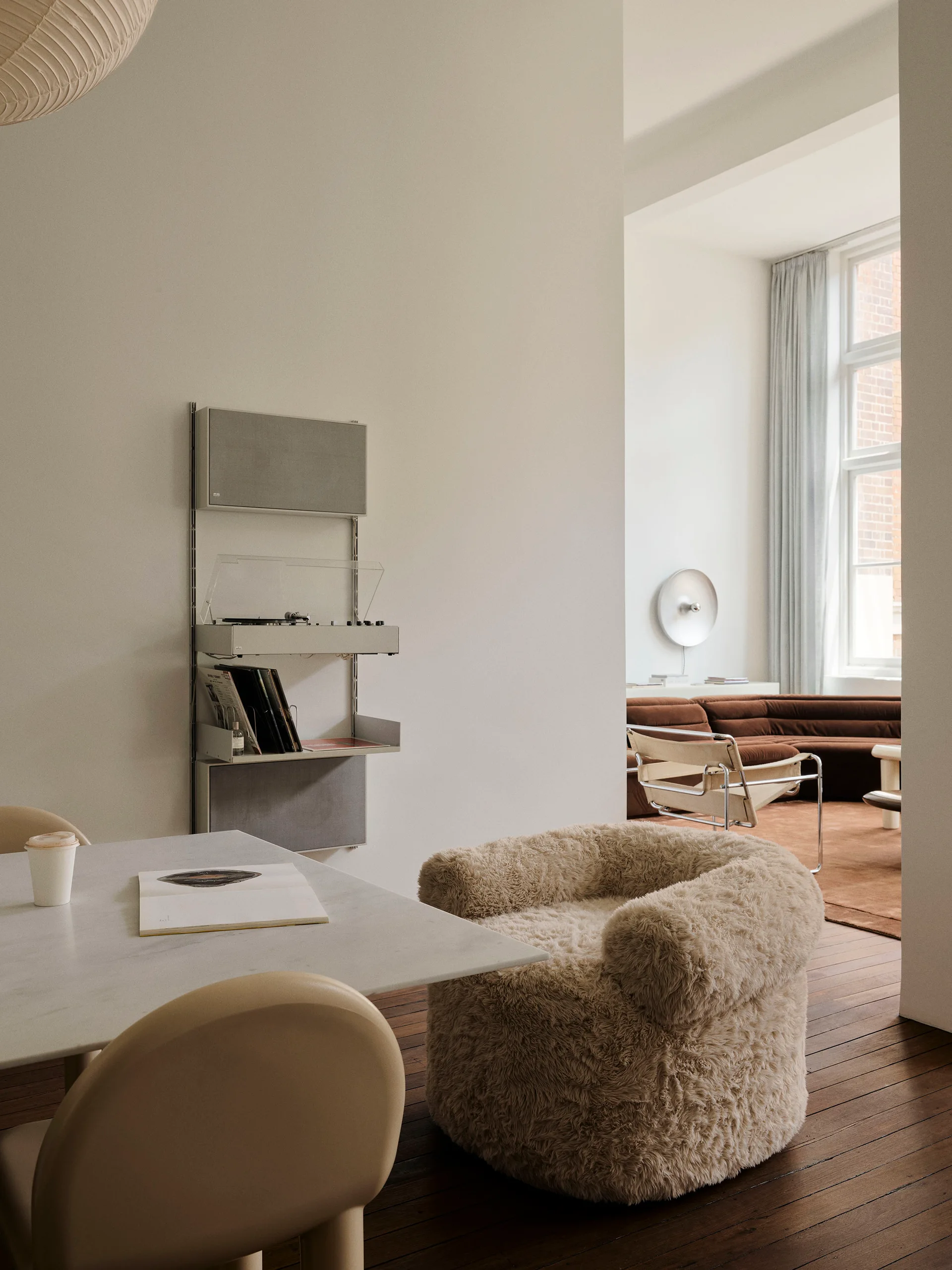 A furry armchair and dining table sit near a modular wall shelf, with a curved brown sofa visible through an opening in the background.