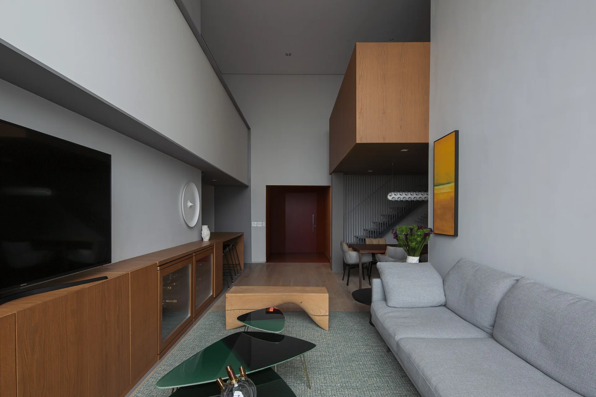 A high-ceilinged grey living room featuring a long wooden media console, a grey sofa, tiered coffee tables, and a suspended wooden loft structure.