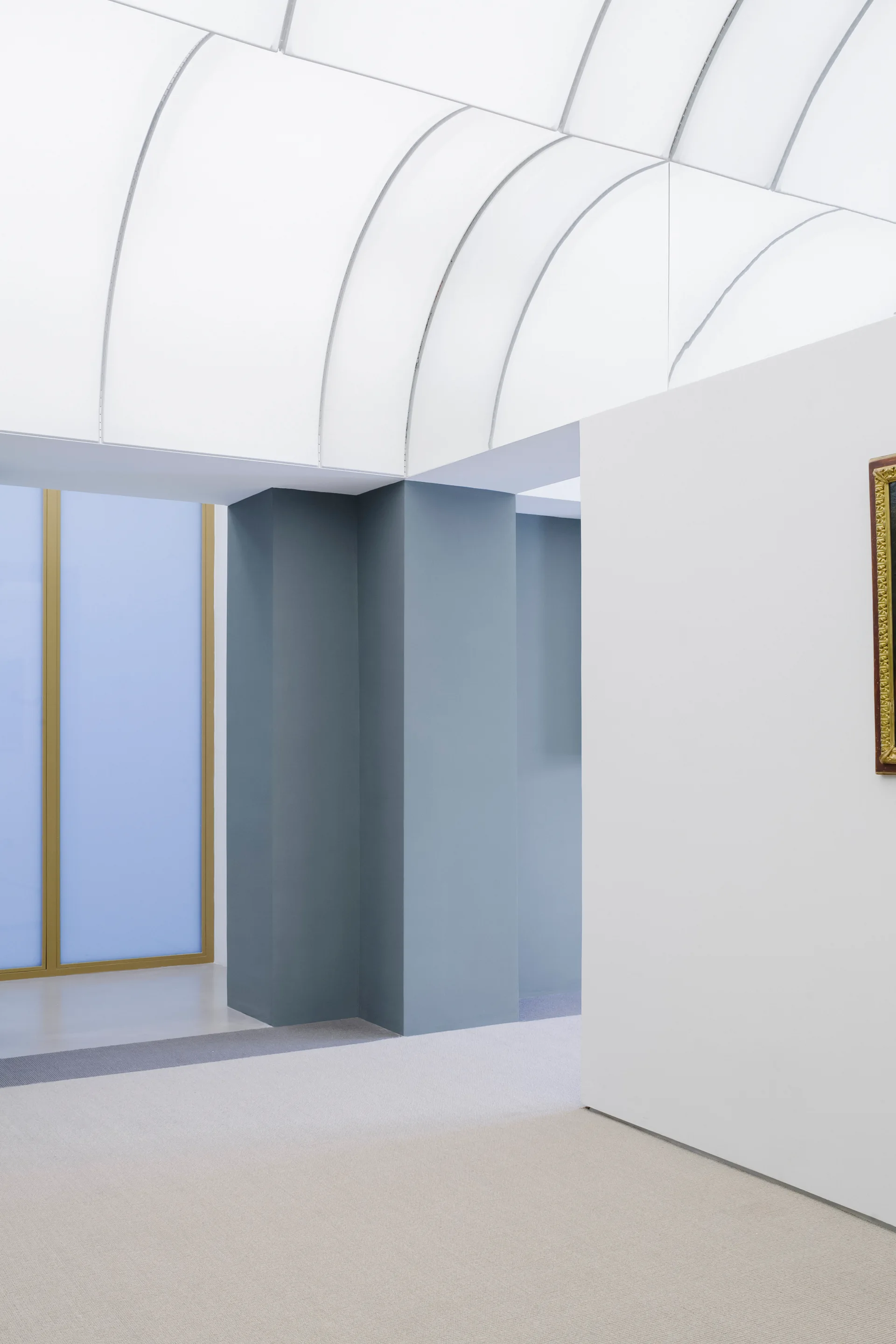A minimalist hallway features a white vaulted ceiling, slate blue structural columns, a grey carpeted floor, and a gold-framed window.