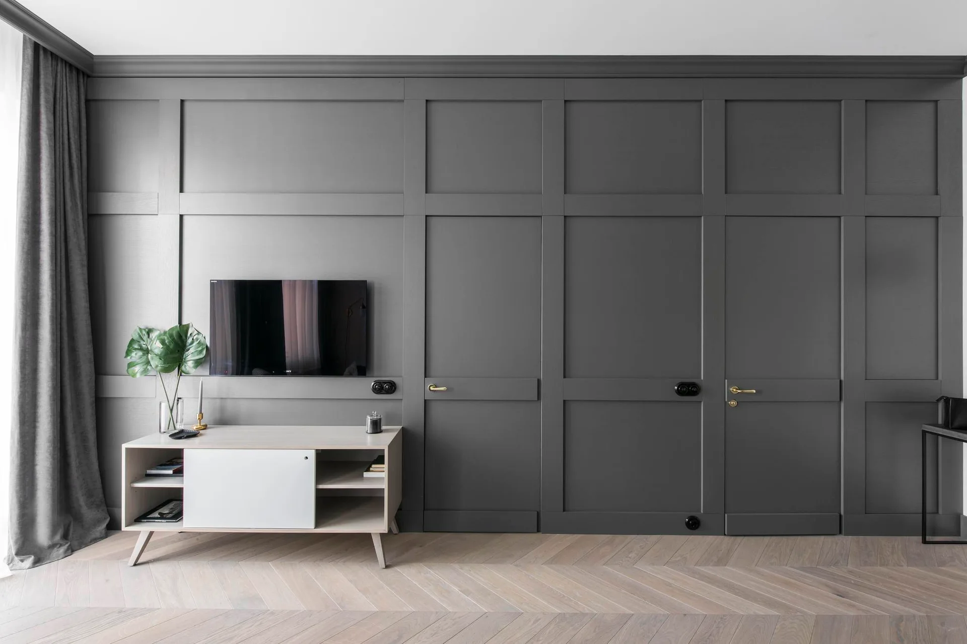 A modern living room features a dark grey paneled wall with integrated doors, a light wood media console, a wall-mounted TV, and herringbone wood flooring.