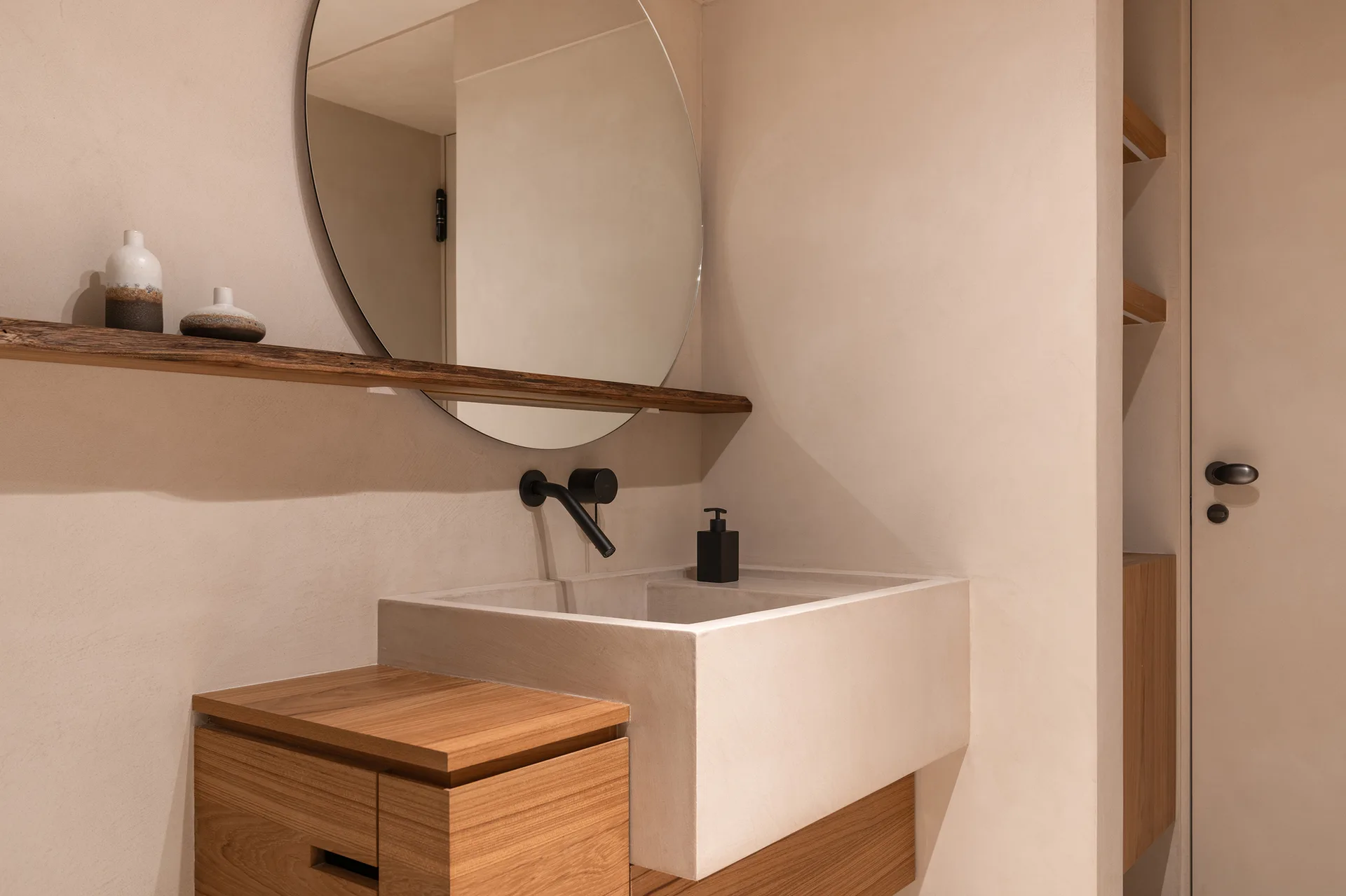 A minimalist bathroom vanity featuring an integrated concrete sink, wall-mounted black faucet, round mirror, and natural wood shelf against plaster walls.