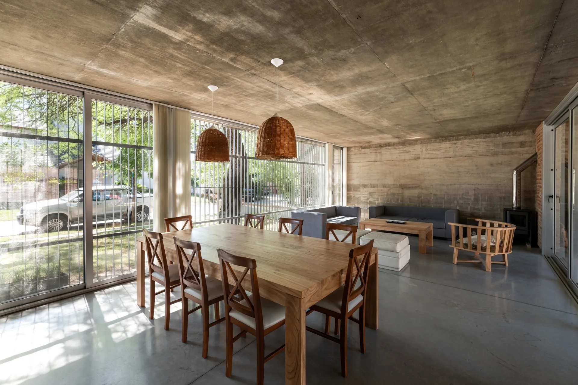 An open dining and living area features a wooden table with chairs under woven pendant lights, with concrete walls and floor-to-ceiling windows.