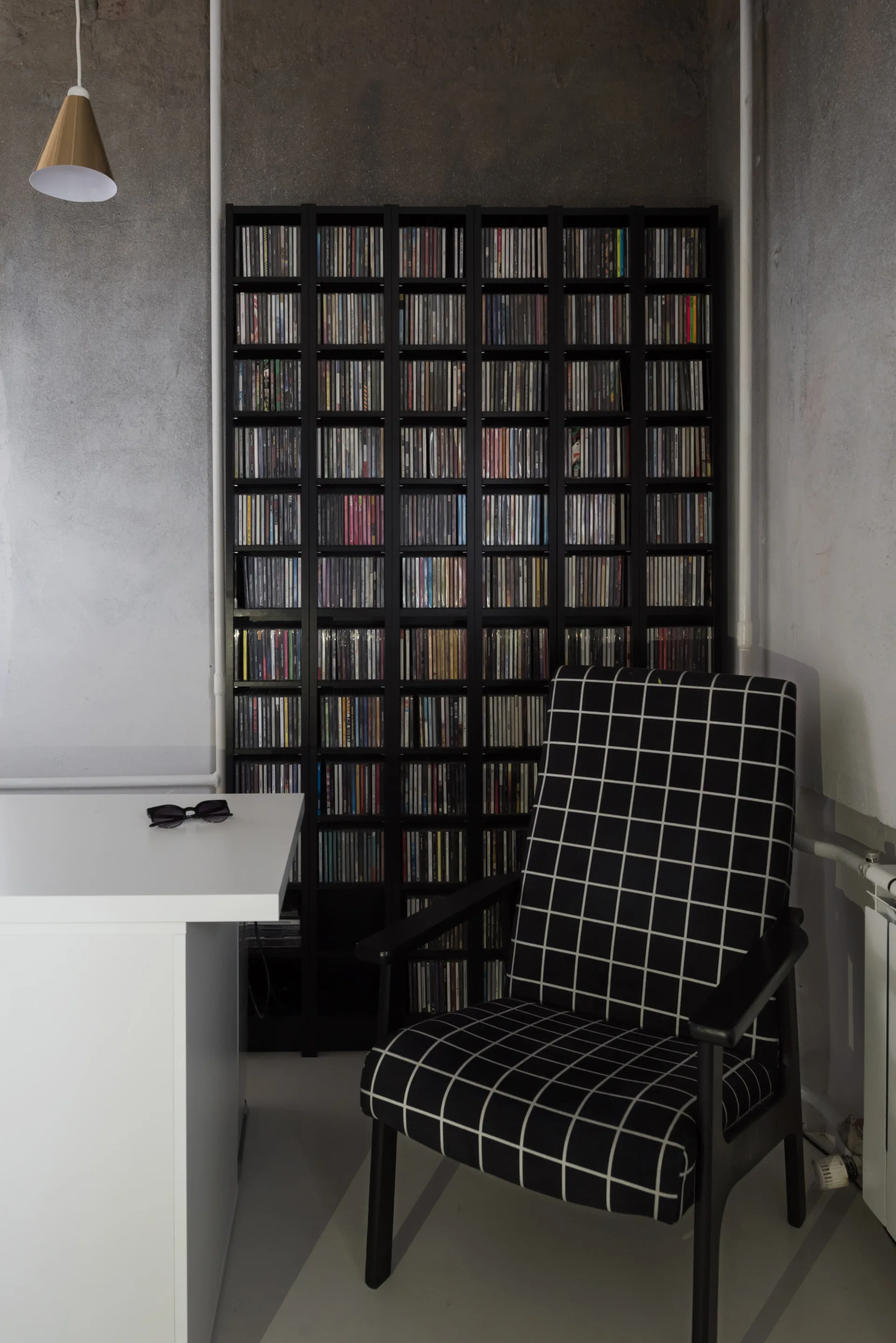 A home office corner featuring a black grid armchair, a white desk, and a tall black media shelf filled with CDs against a textured gray wall.