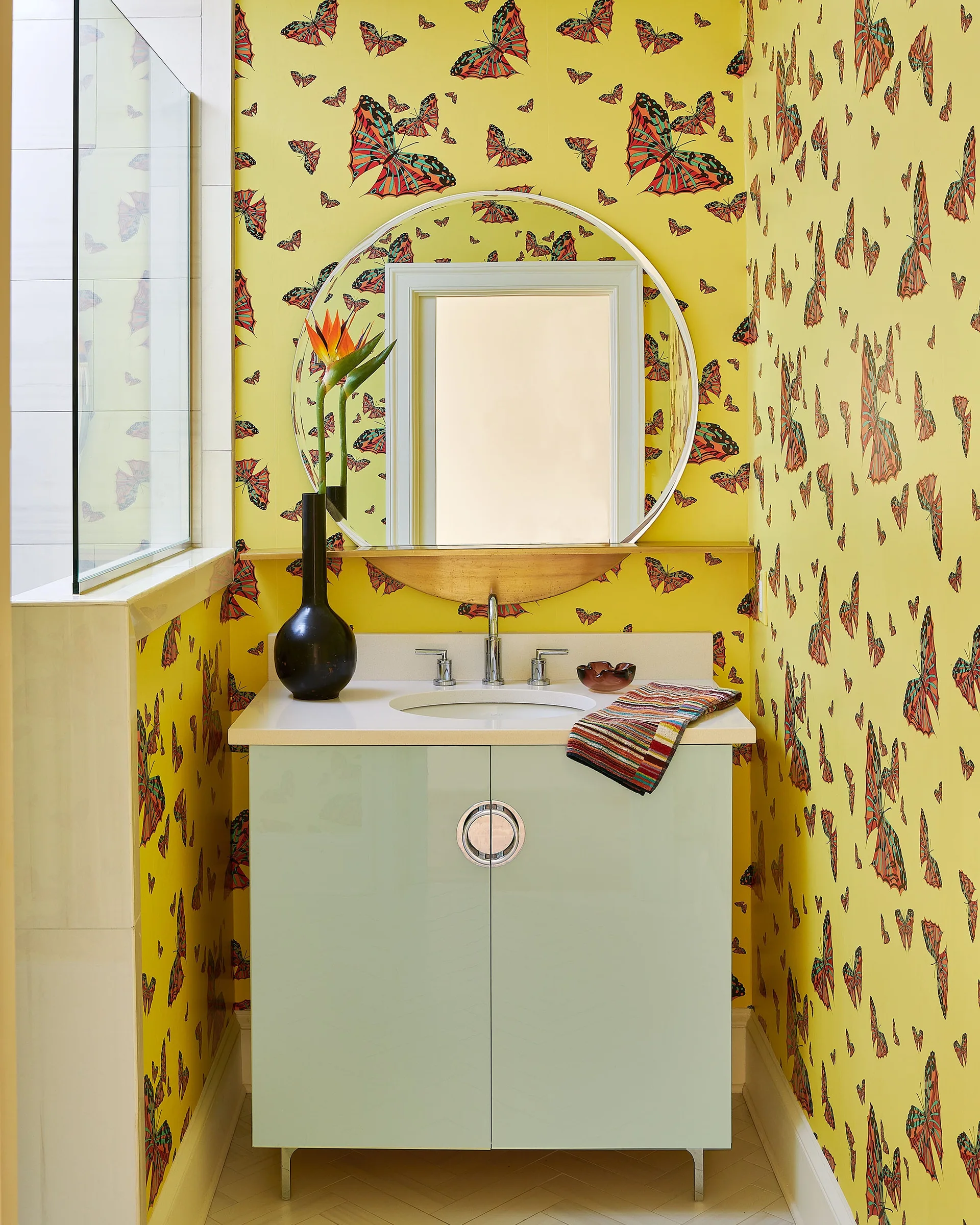 A small powder room features vibrant yellow butterfly wallpaper, a pale green gloss vanity with a round mirror, and a black vase.