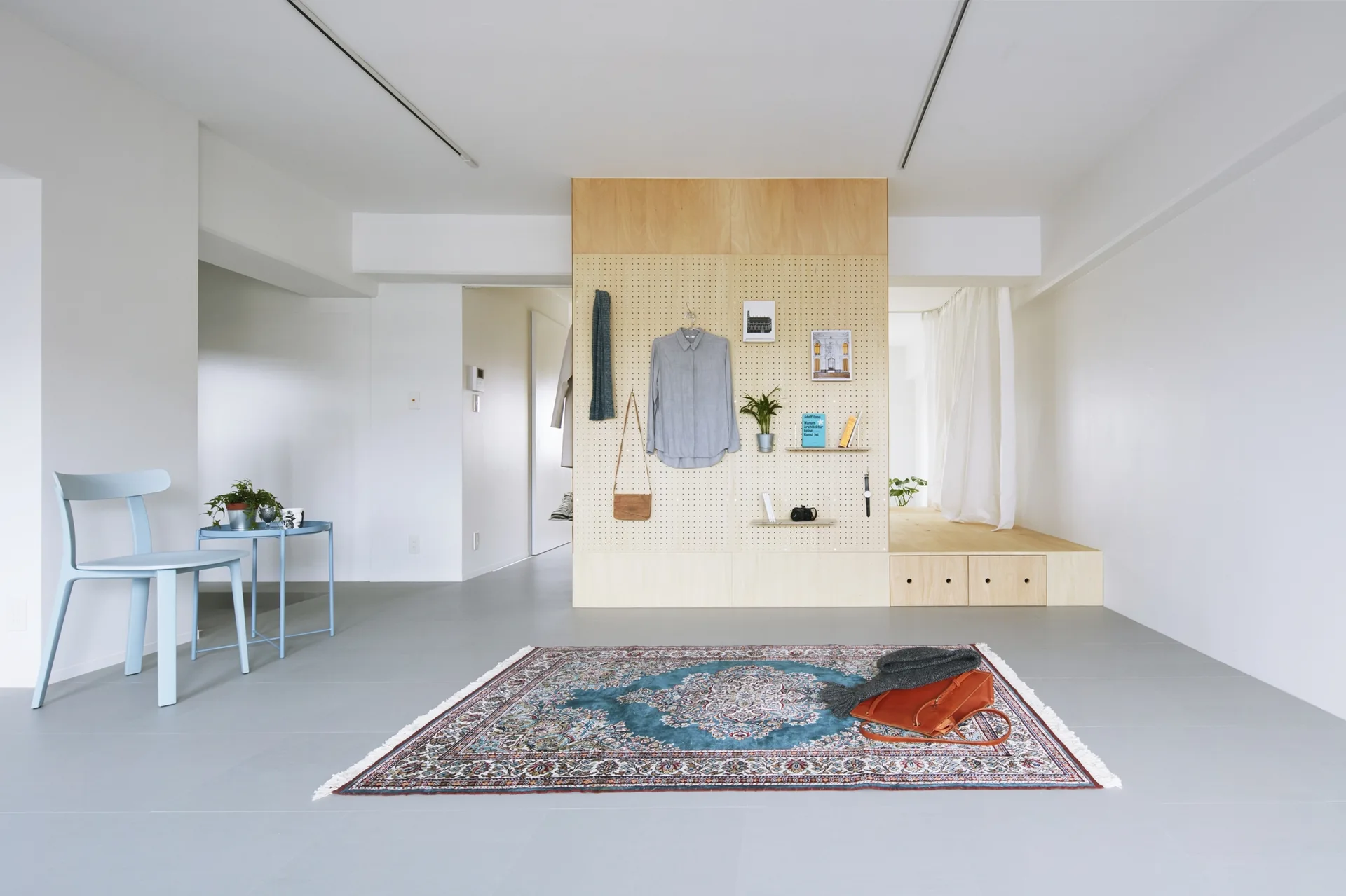 A minimalist living room features a wooden pegboard room divider, a raised wooden platform, a light blue chair, and an ornate rug on a grey floor.