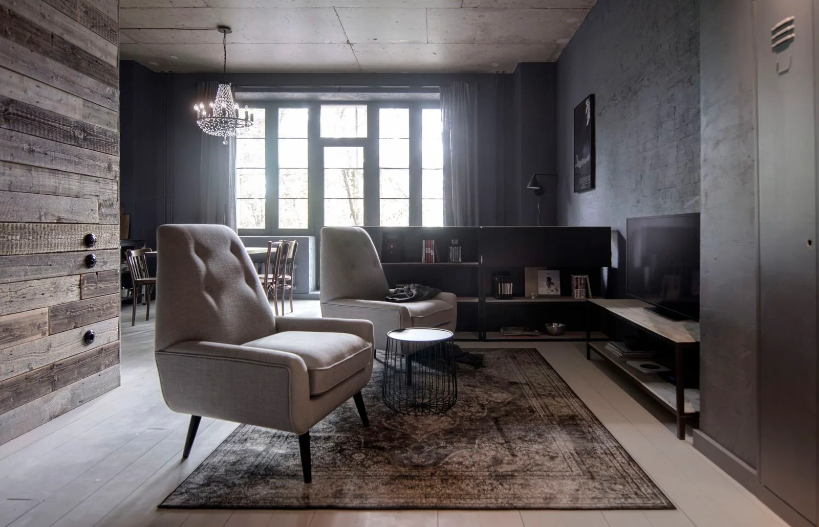 Two grey armchairs sit on a distressed rug in an industrial living room with a concrete ceiling and dark grey walls.