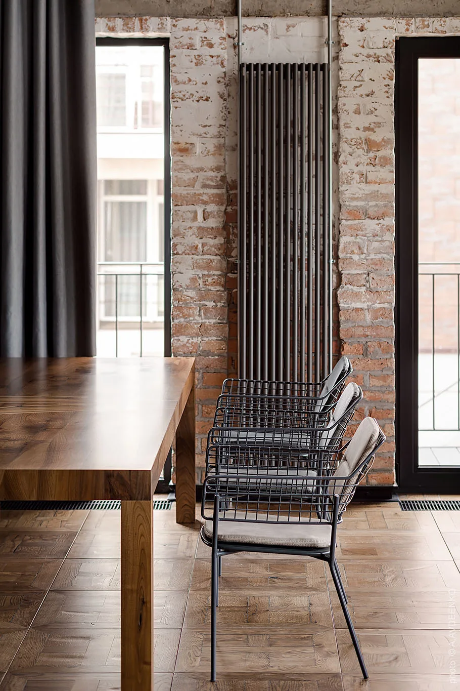 A solid wood dining table sits next to a row of grey wire chairs against an exposed brick wall with tall windows.