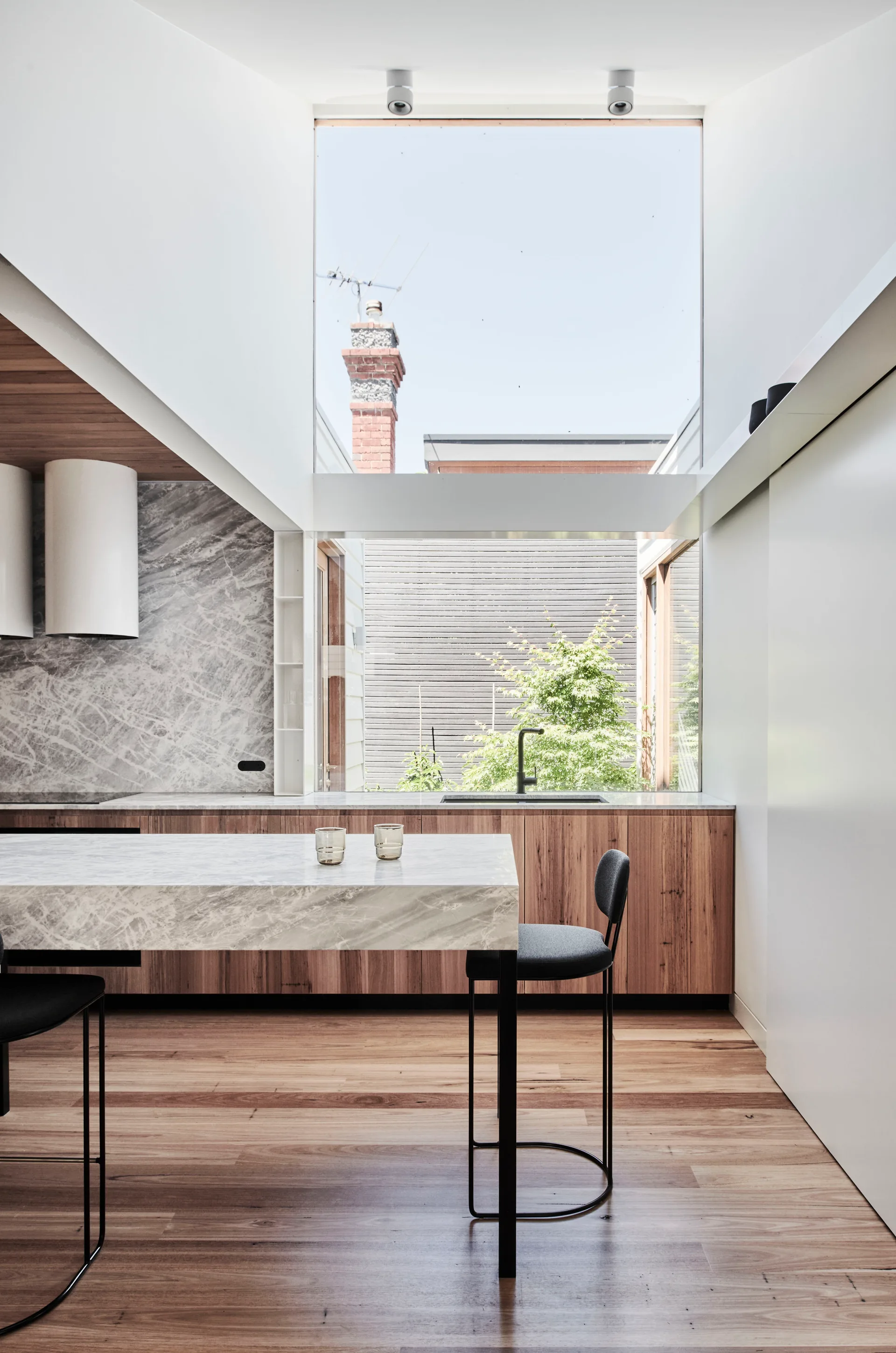 A contemporary kitchen features a marble-topped island with black bar stools, wood cabinetry, and a soaring skylight window overlooking the sky.