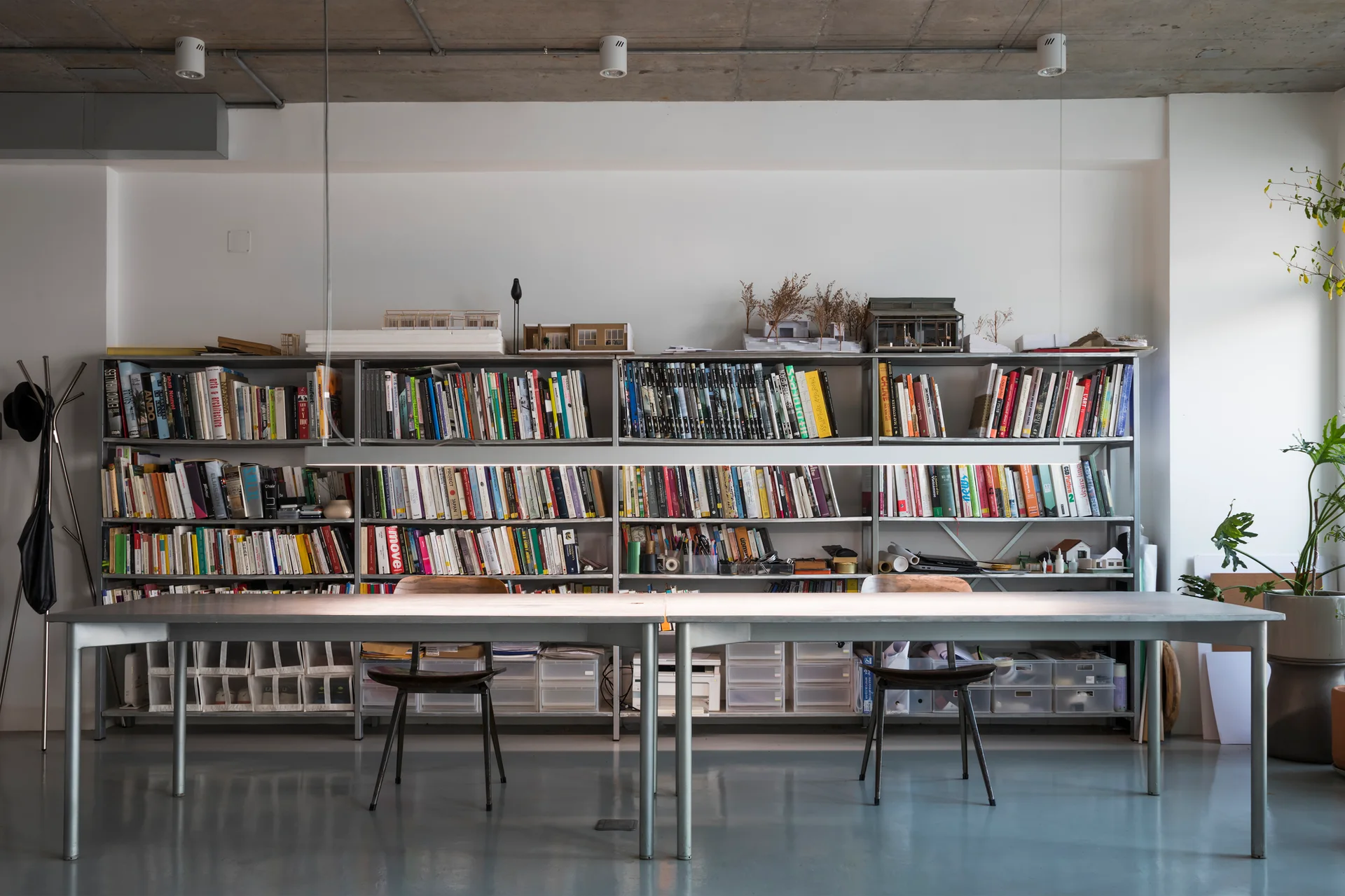 A minimalist studio workspace featuring a long metal table, wooden chairs, and a large metal bookshelf filled with books and architectural models.