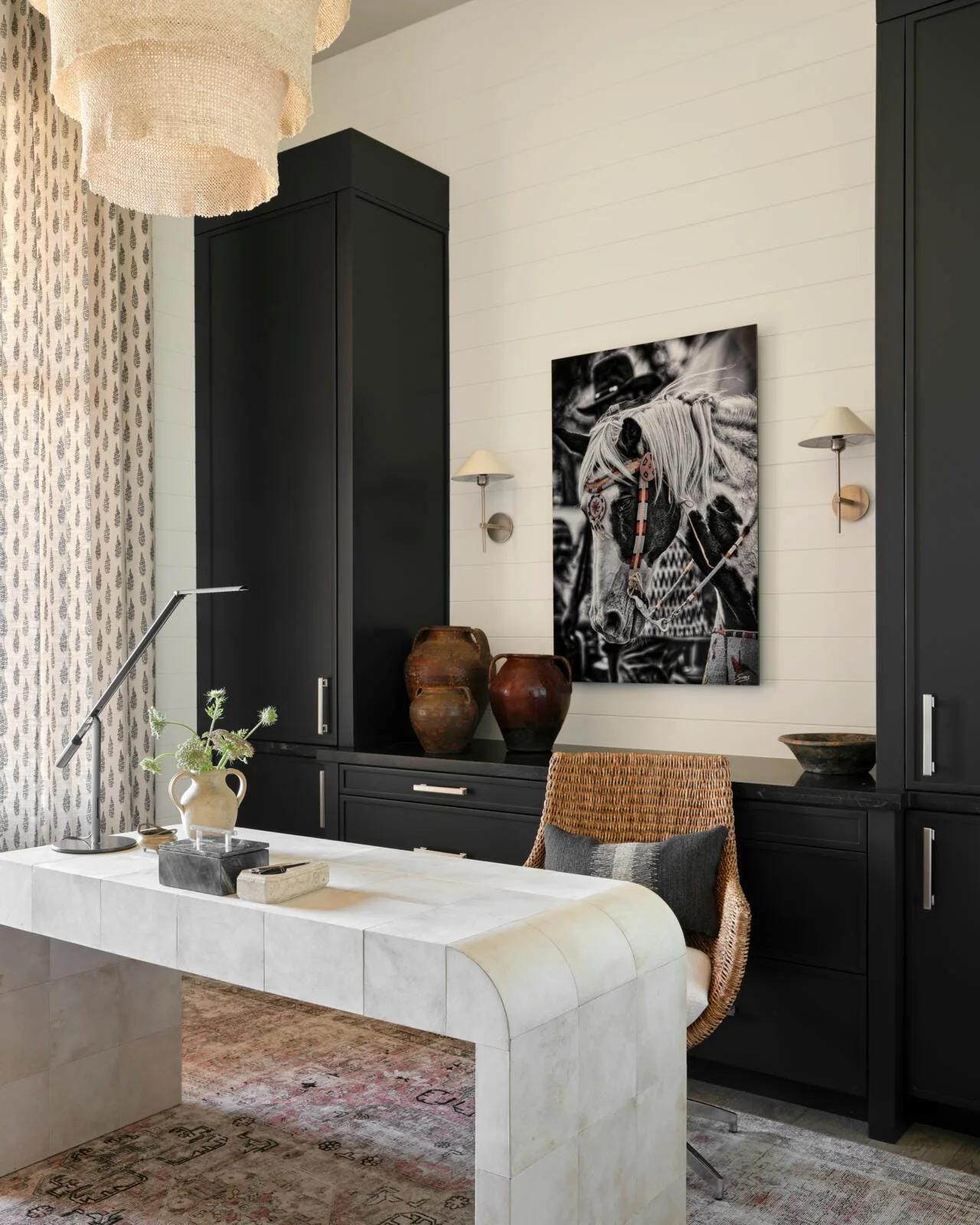 A home office featuring a light-colored waterfall desk, a woven rattan chair, and dark black floor-to-ceiling built-in cabinets on a shiplap wall.