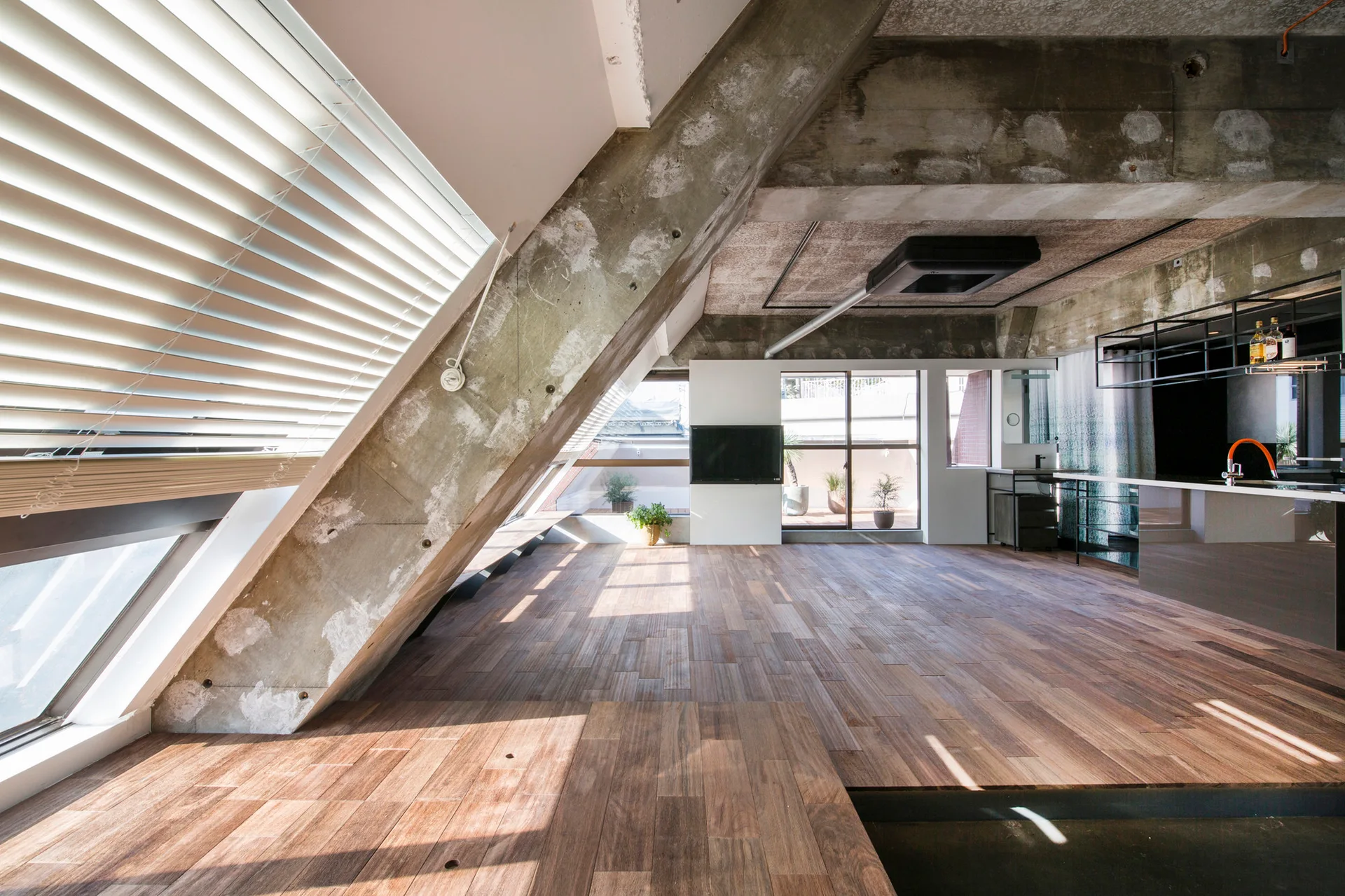 An open industrial loft features slanted raw concrete columns, hardwood flooring, a modern kitchen island, and sunlight filtering through sloped window blinds.