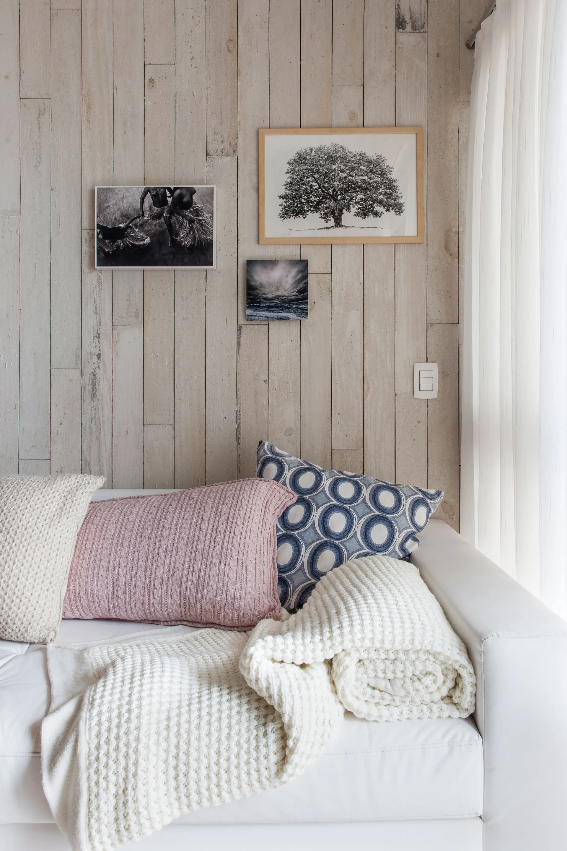 A white sofa is dressed with knit throw pillows and a blanket against a whitewashed vertical wood plank wall decorated with three small framed artworks.