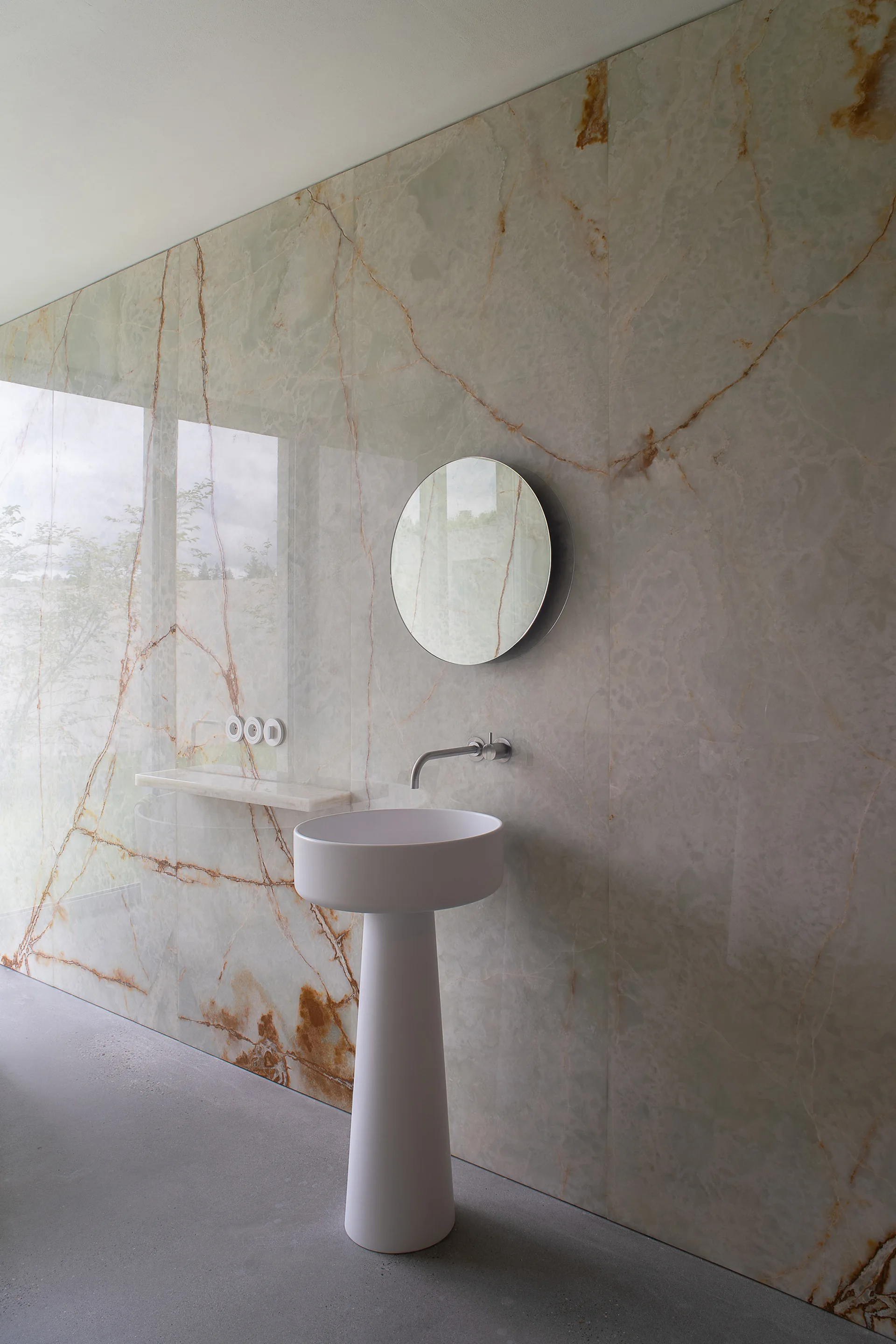 A minimalist bathroom features a white pedestal sink, a wall-mounted faucet, a round mirror, and a large-scale veined green stone wall.