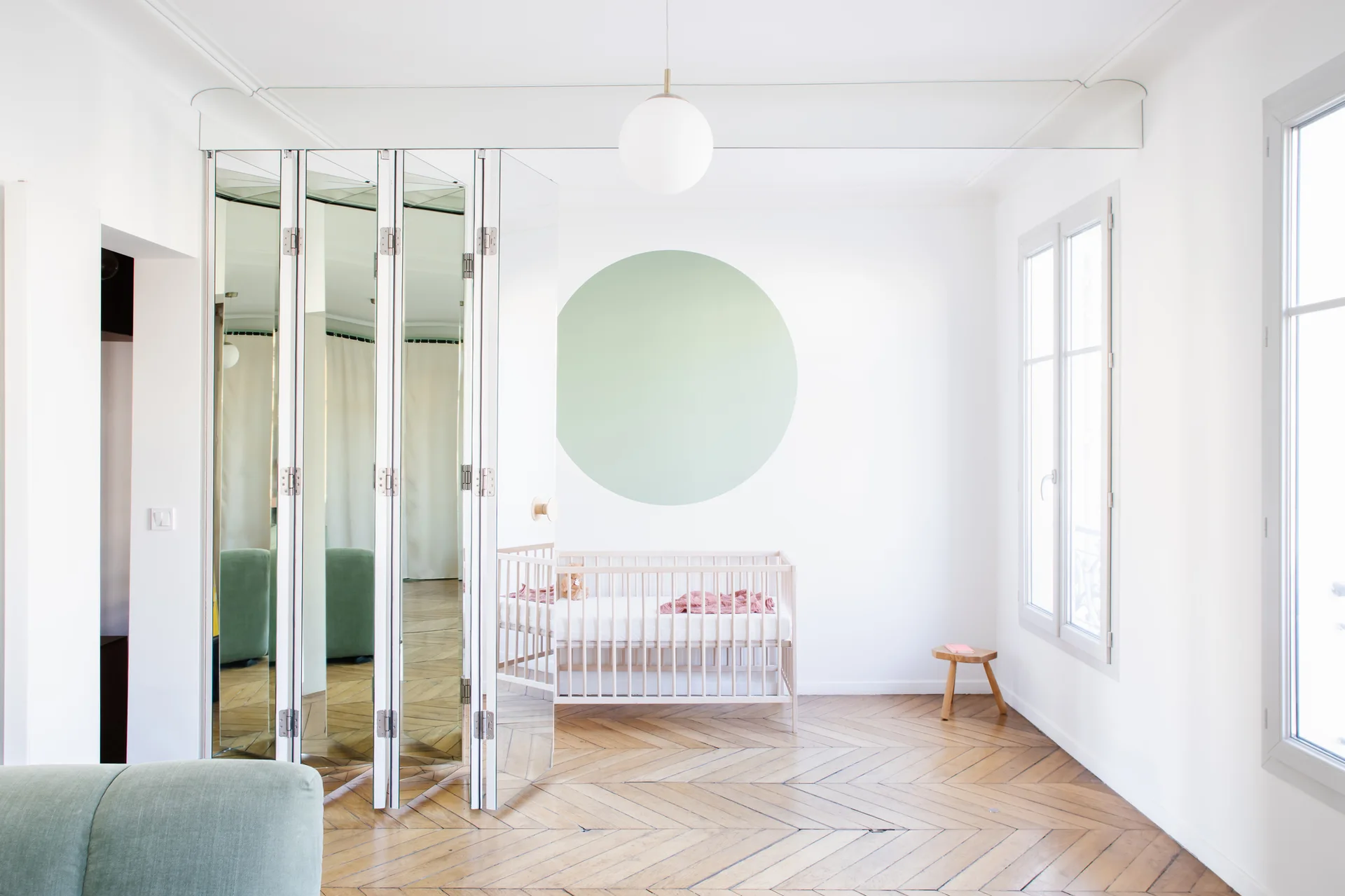 A bright nursery featuring a light wood crib under a painted green circle next to tall white folding mirrored doors and herringbone floors.