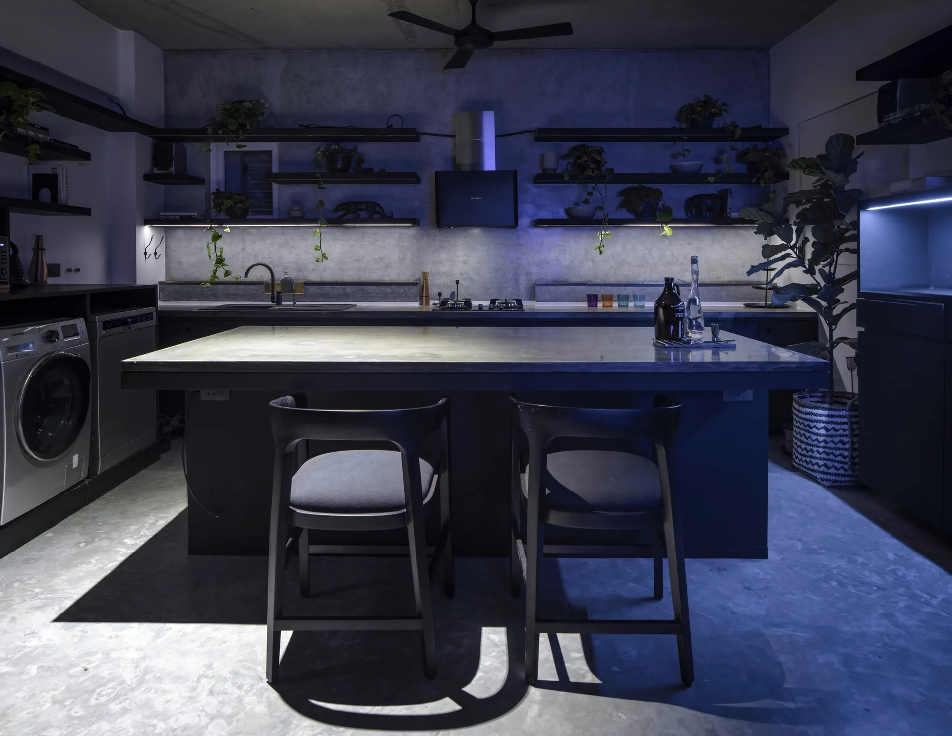 A dark industrial kitchen with a concrete backsplash, floating shelves with plants, a large black island with two chairs, and cool blue ambient lighting.
