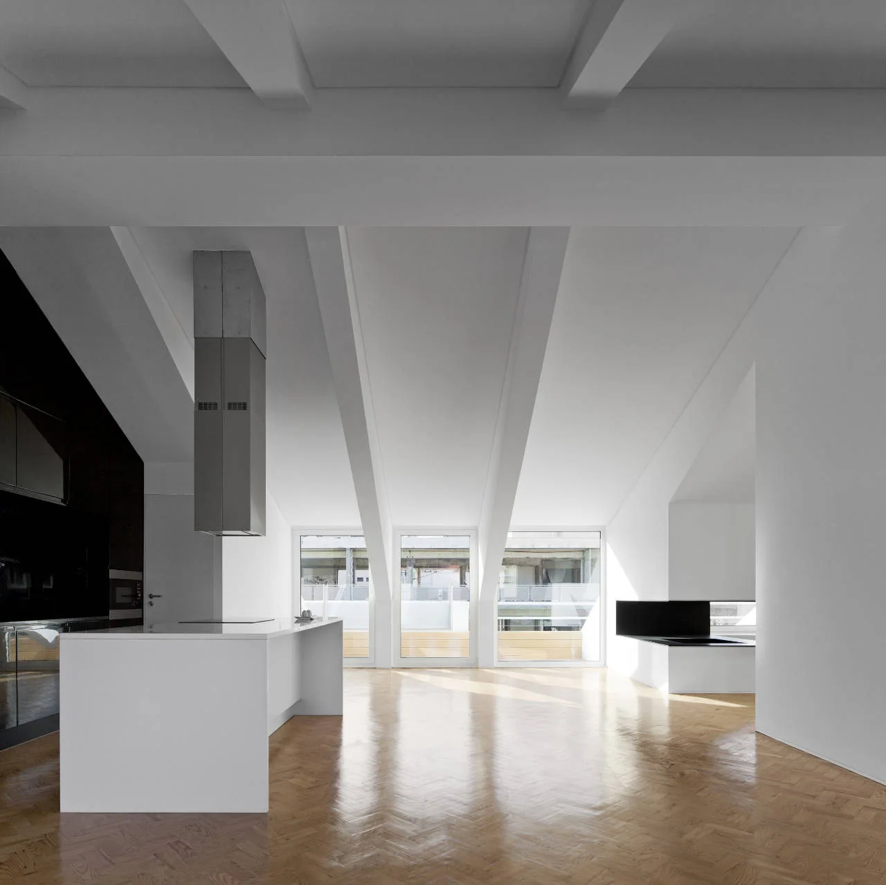 A minimalist kitchen features a white island, dark cabinetry, and herringbone wood floors beneath a slanted white ceiling with structural beams.