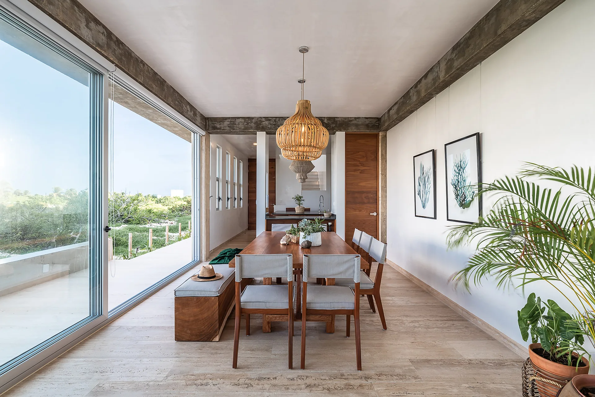 A coastal dining room features a long wooden table with light gray chairs, a low bench, a wooden pendant light, and a massive sliding glass door.