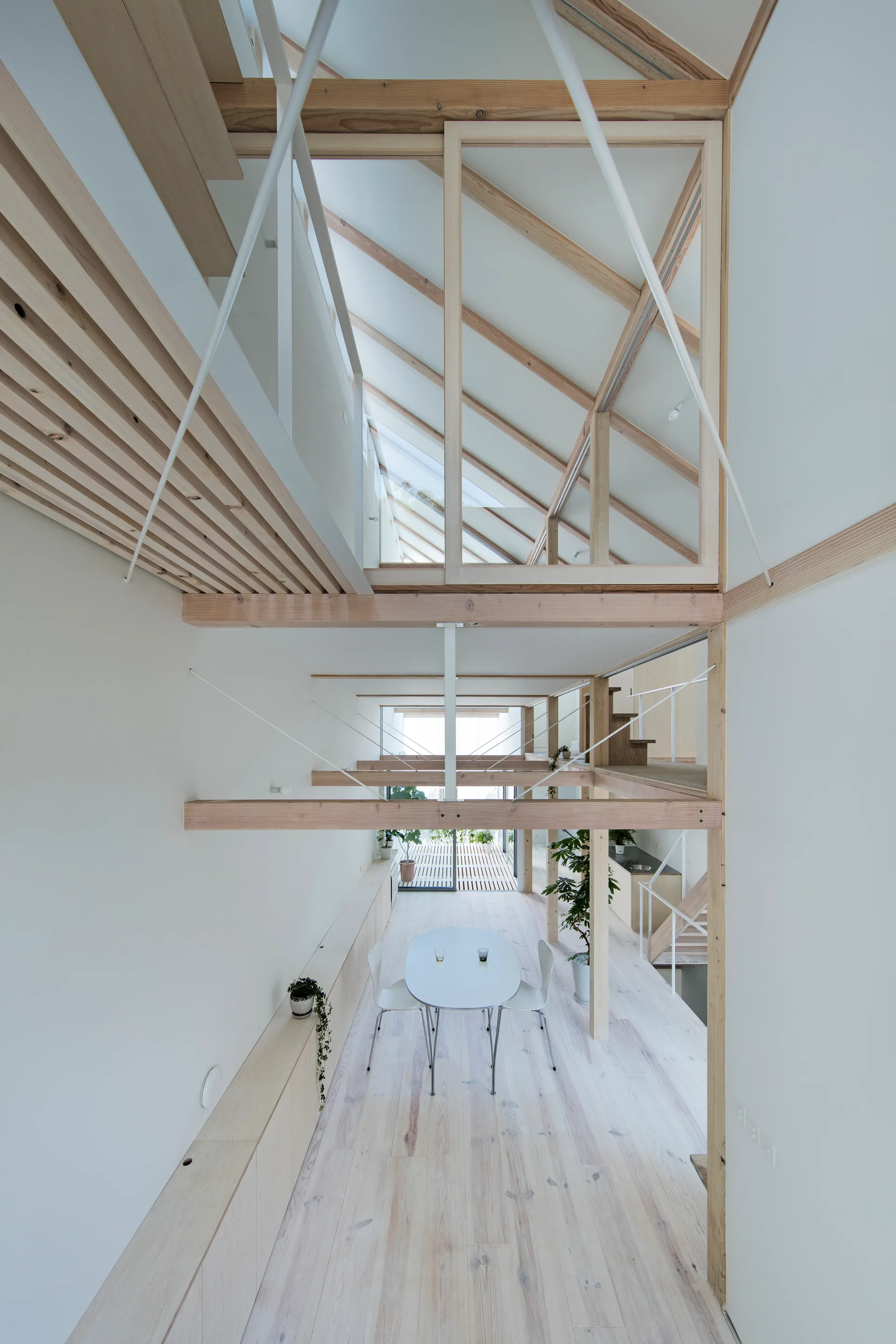 A high-angle view of a minimalist open-plan home featuring a light wood dining set, white walls, and exposed timber rafters on a vaulted ceiling.