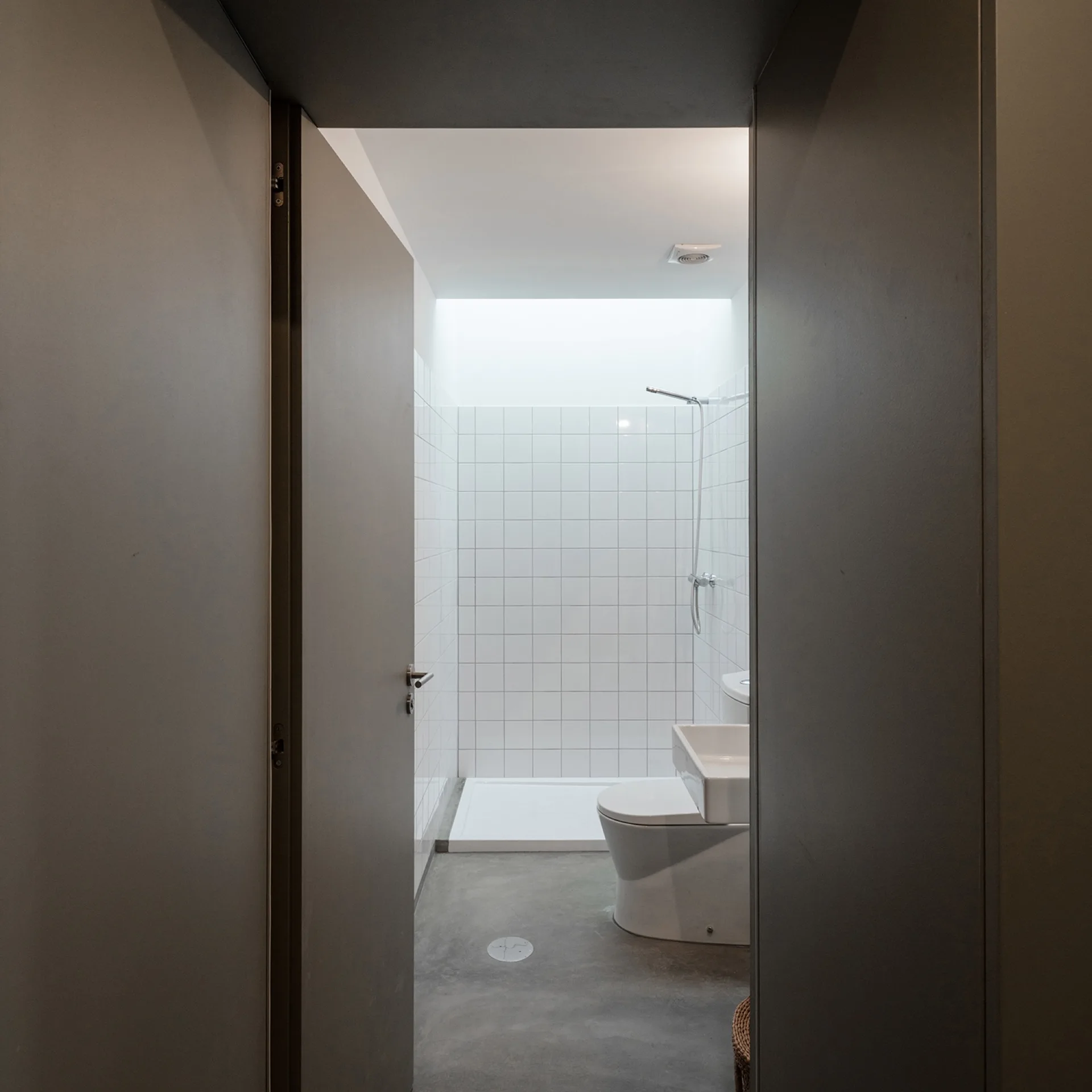 A view through a dark doorway into a minimalist bathroom with white square wall tiles, a toilet, and grey concrete flooring.