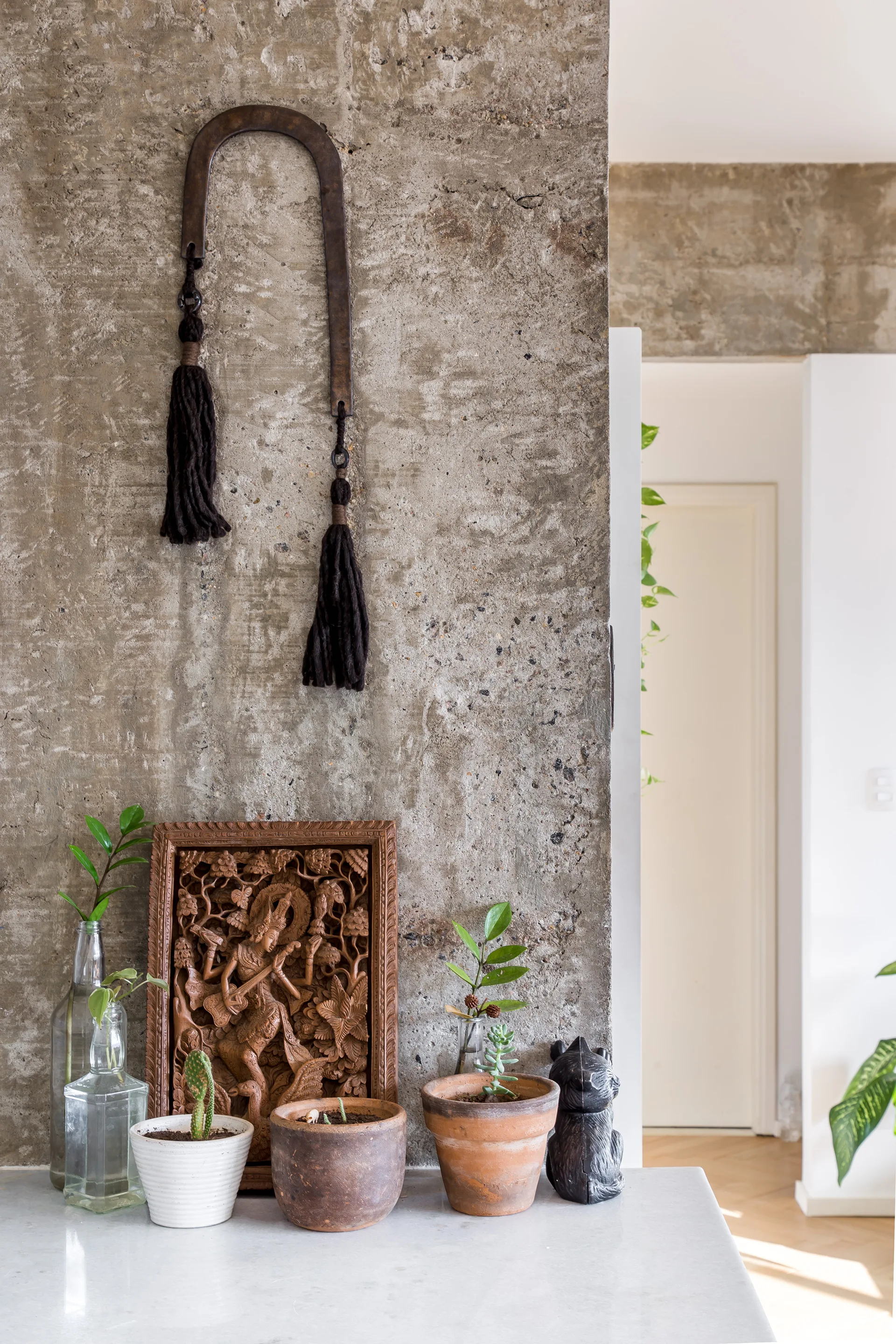 A textured raw concrete wall is decorated with a carved wood panel, hanging metal horseshoe with tassels, and small potted plants on a white counter.