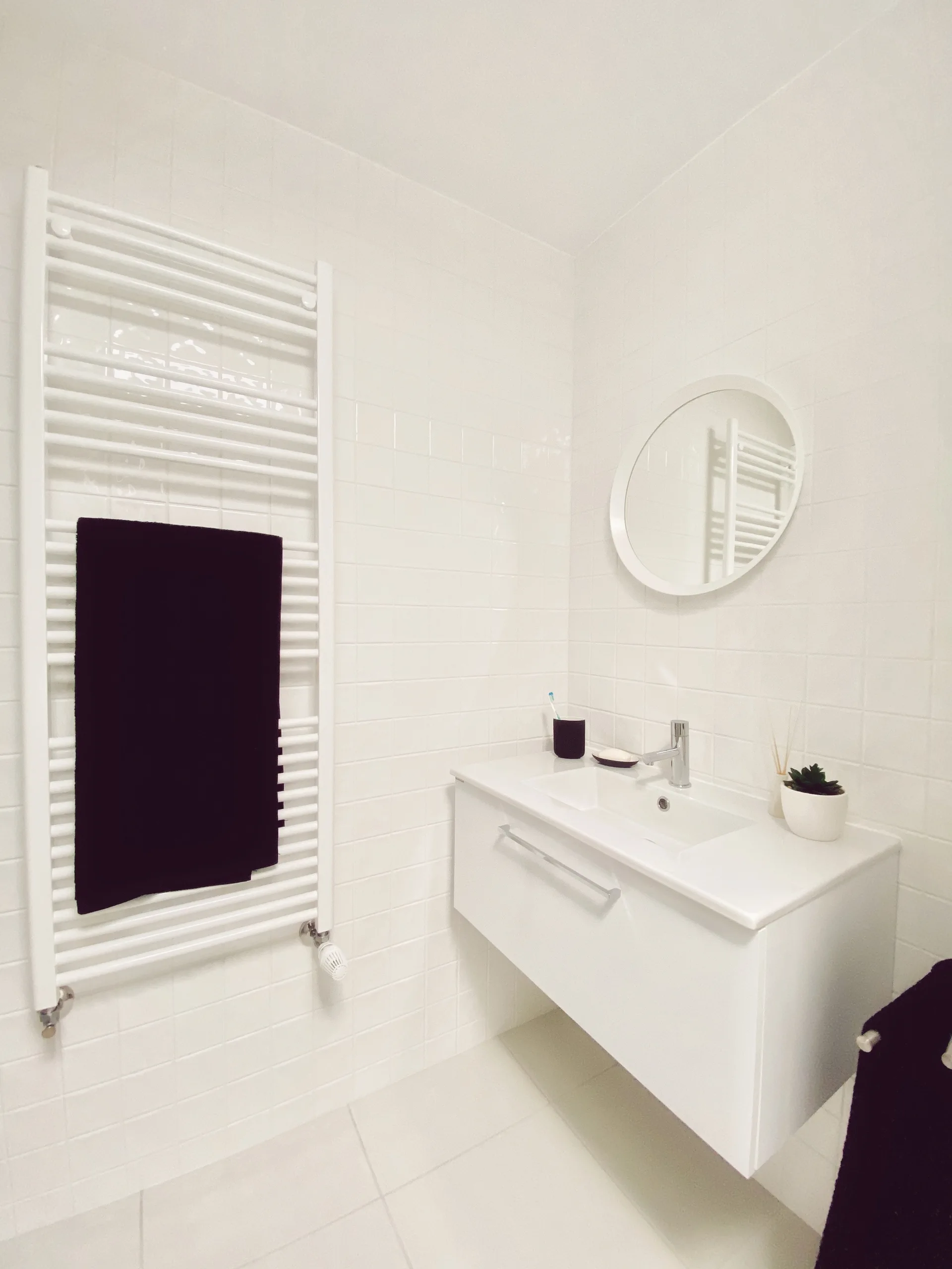 A minimalist white tiled bathroom featuring a wall-mounted floating vanity, a round mirror, and a white ladder towel warmer holding a black towel.