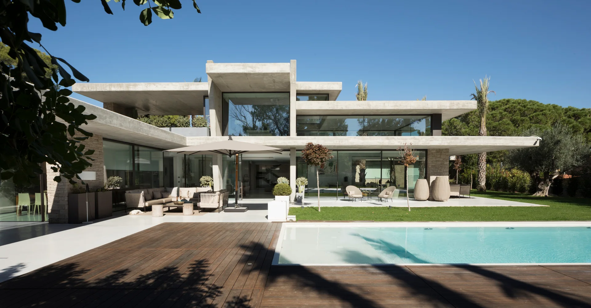 A modern concrete residence with large glass windows, an outdoor patio seating area, and a turquoise pool with dark wood decking.