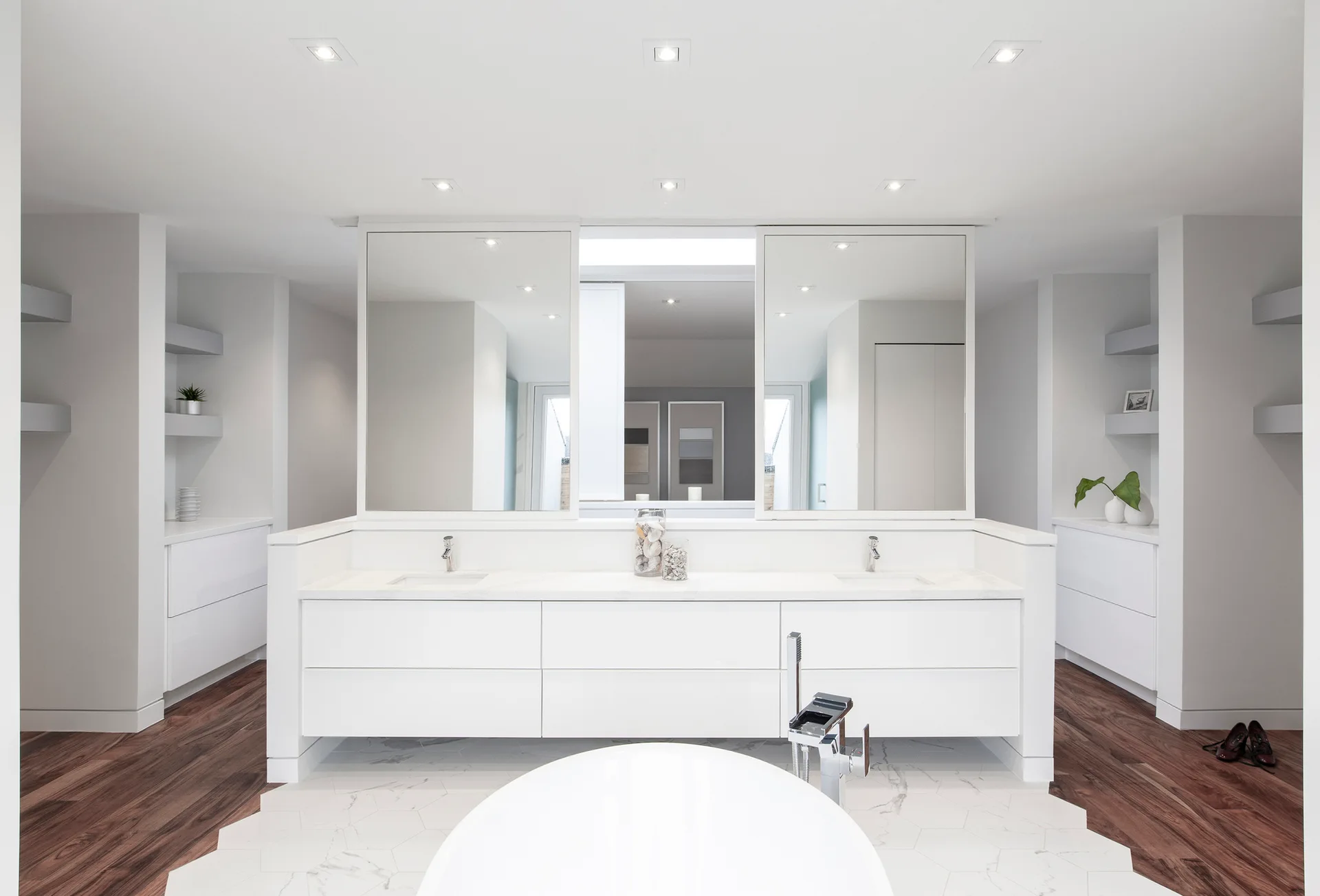 Symmetrical white bathroom featuring a double vanity, a freestanding tub, and a floor transitioning from hexagonal marble tile to dark wood.