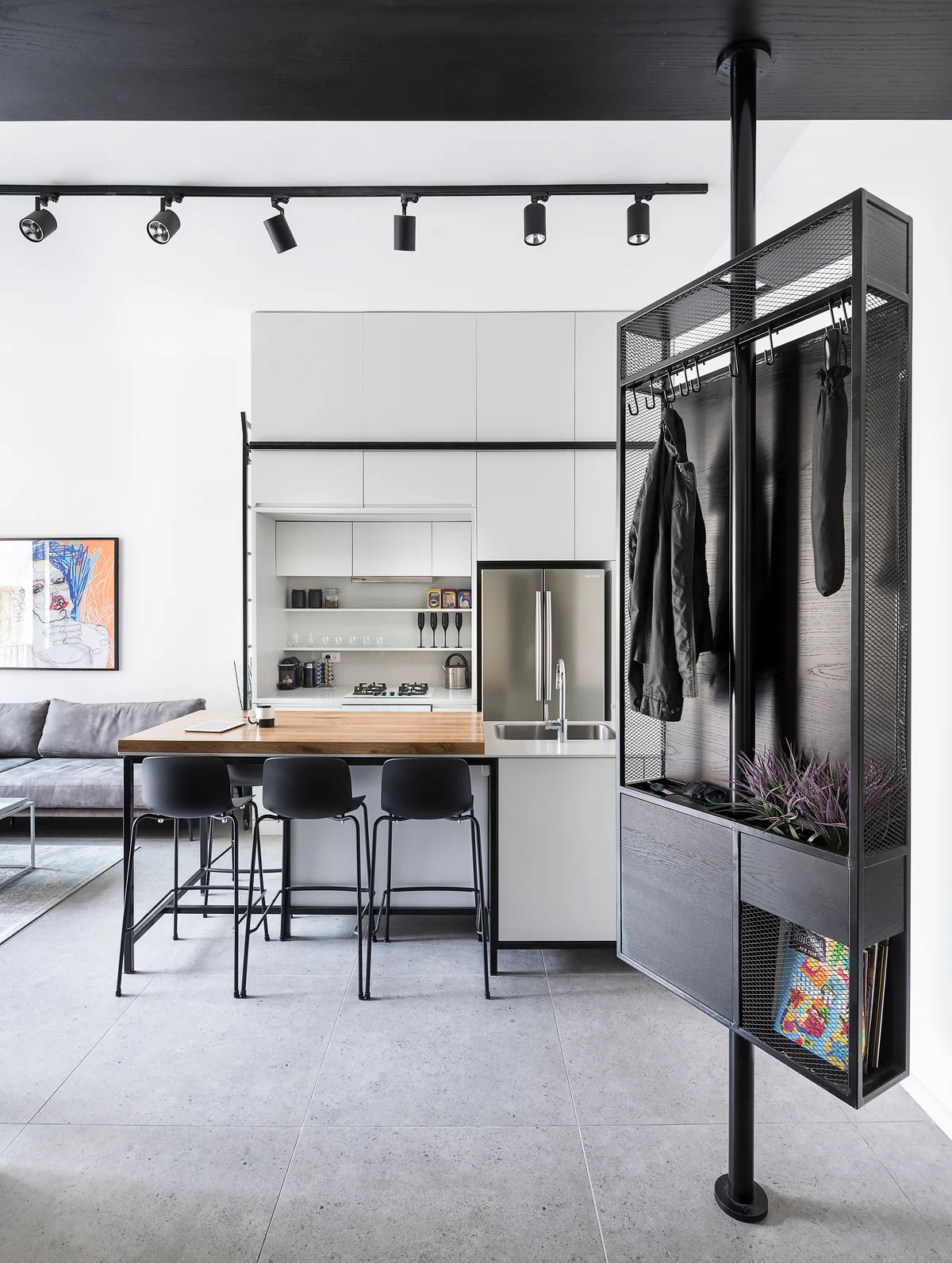 A modern kitchen featuring a wood-topped island with black bar stools, white minimalist cabinetry, and a floor-to-ceiling black metal mesh partition.