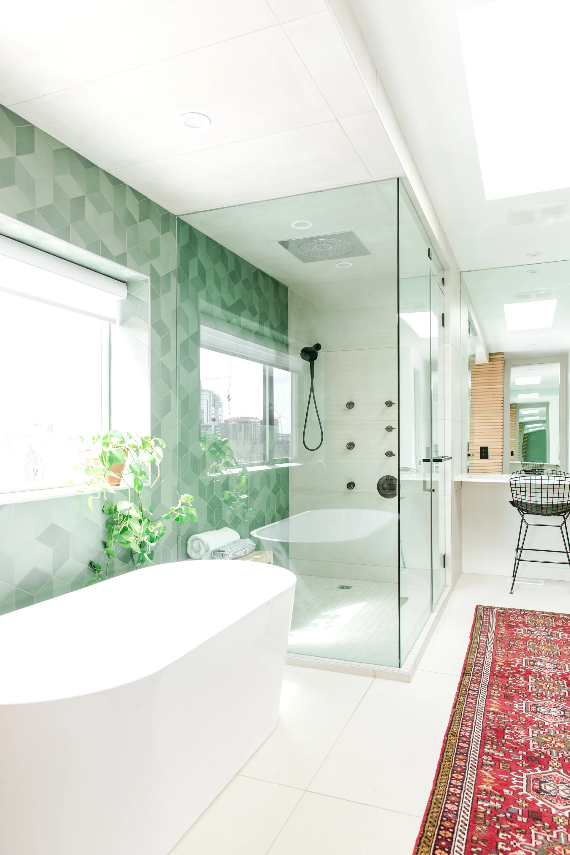 A modern bathroom featuring a white freestanding bathtub, a glass shower with sage green geometric tiles, and a red patterned runner rug.