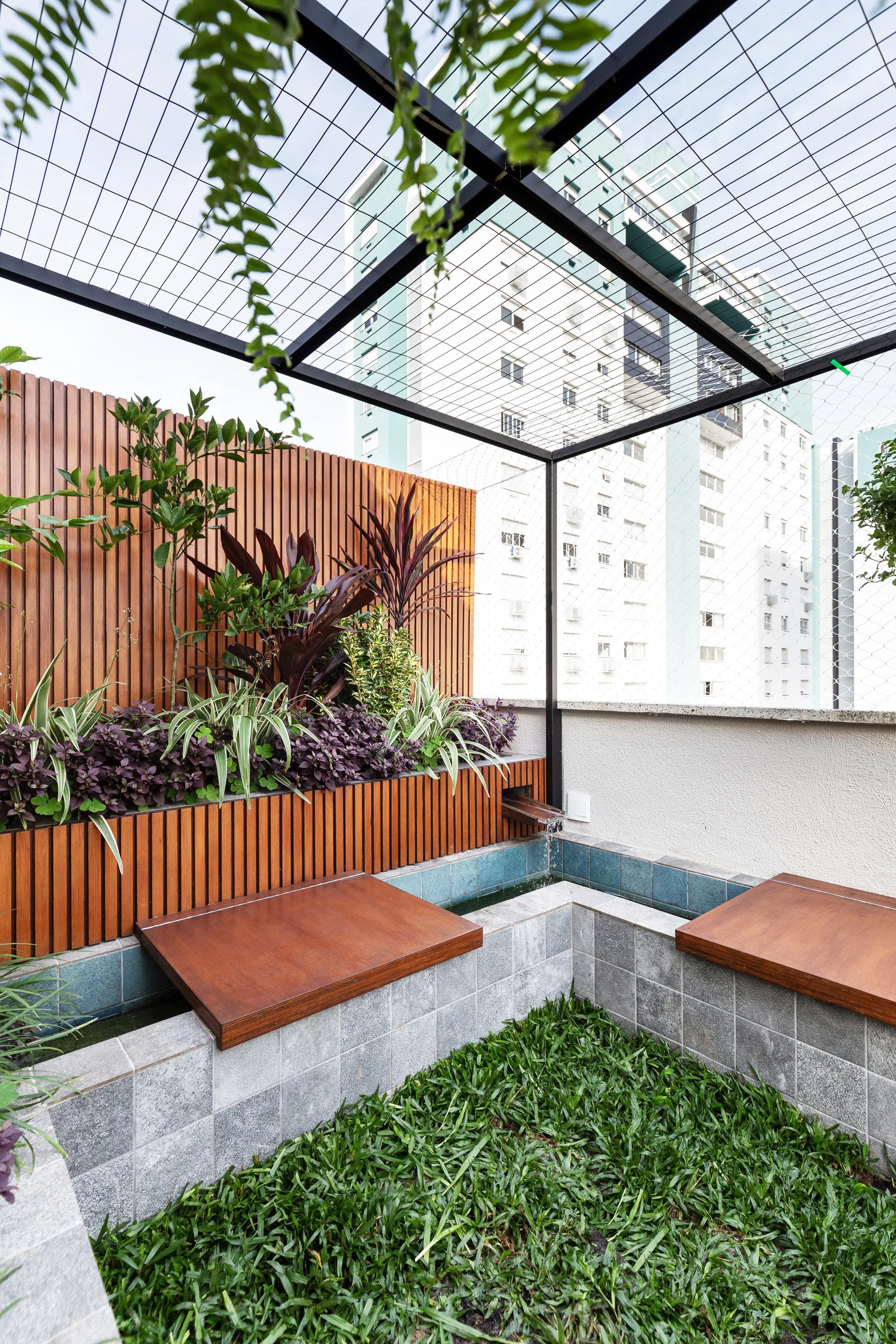An urban rooftop terrace features a vertical wood slat wall, lush planter beds, a stone water basin with blue tiles, and wood-topped benches.
