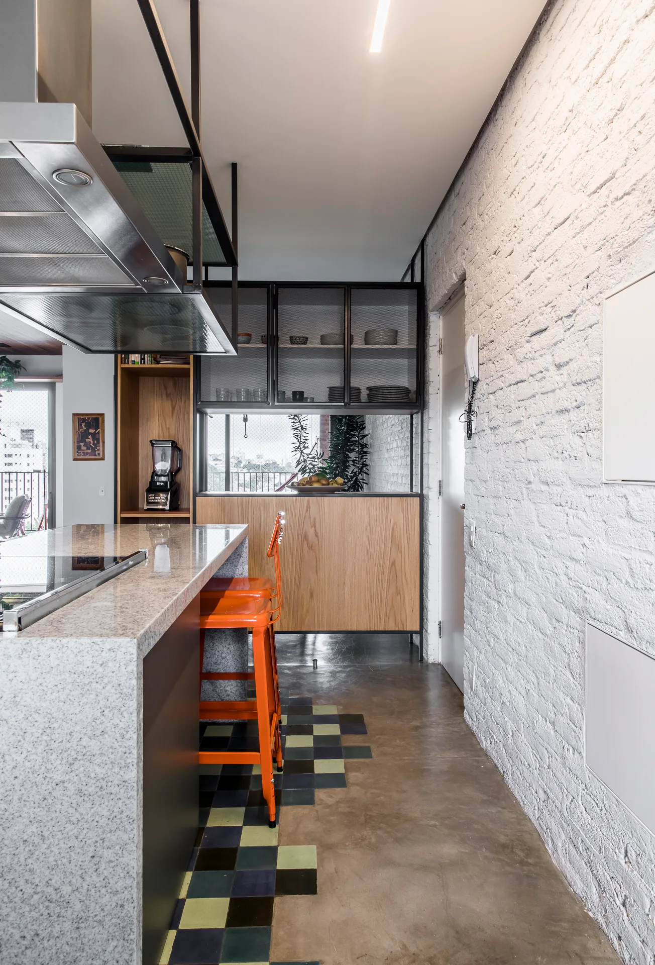 A modern industrial kitchen featuring a stone-topped island with orange metal stools, a white exposed brick wall, and checkered floor tiles.