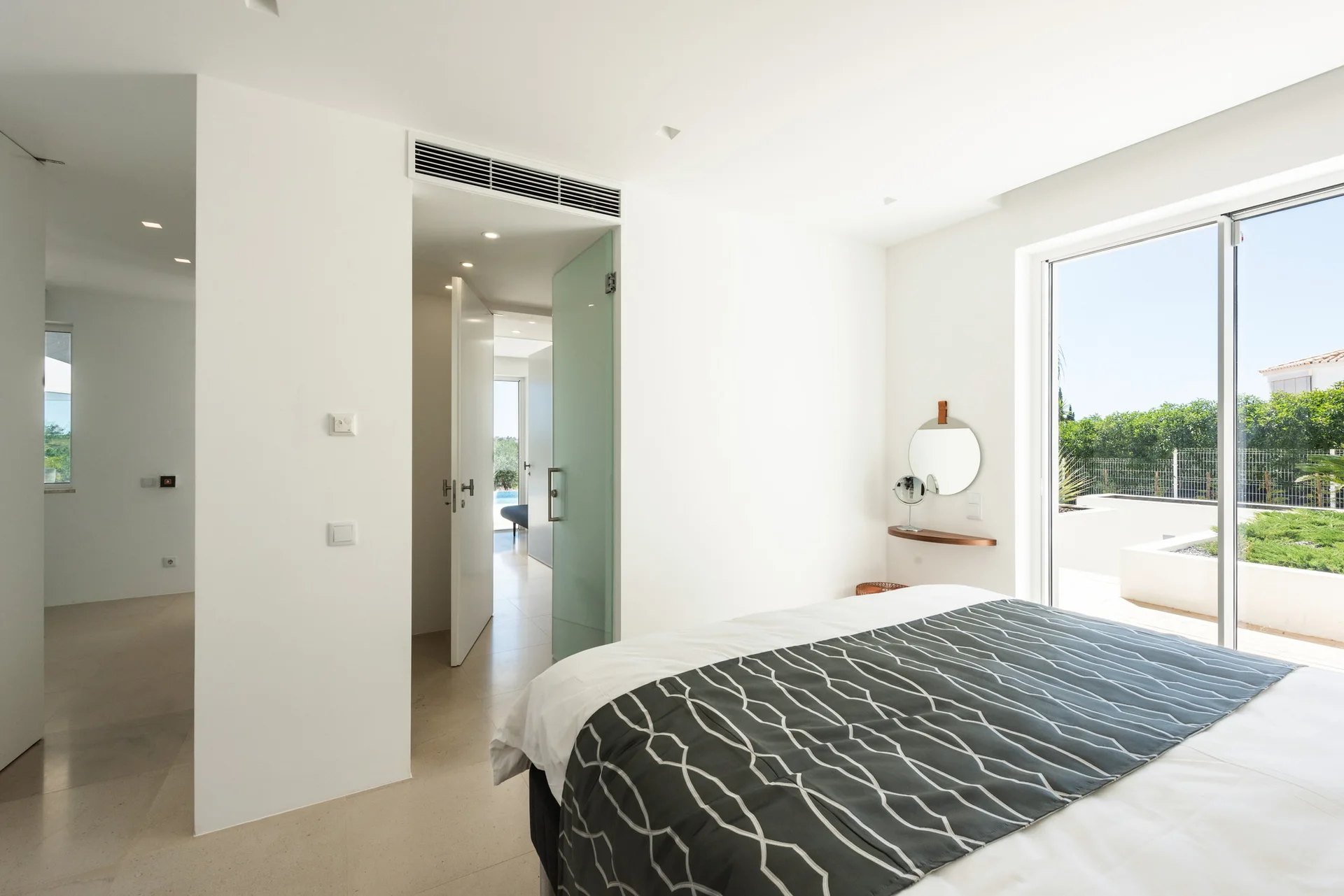 A white minimalist bedroom features a bed with a geometric runner, a small floating wood shelf with a mirror, and a large sliding glass terrace door.