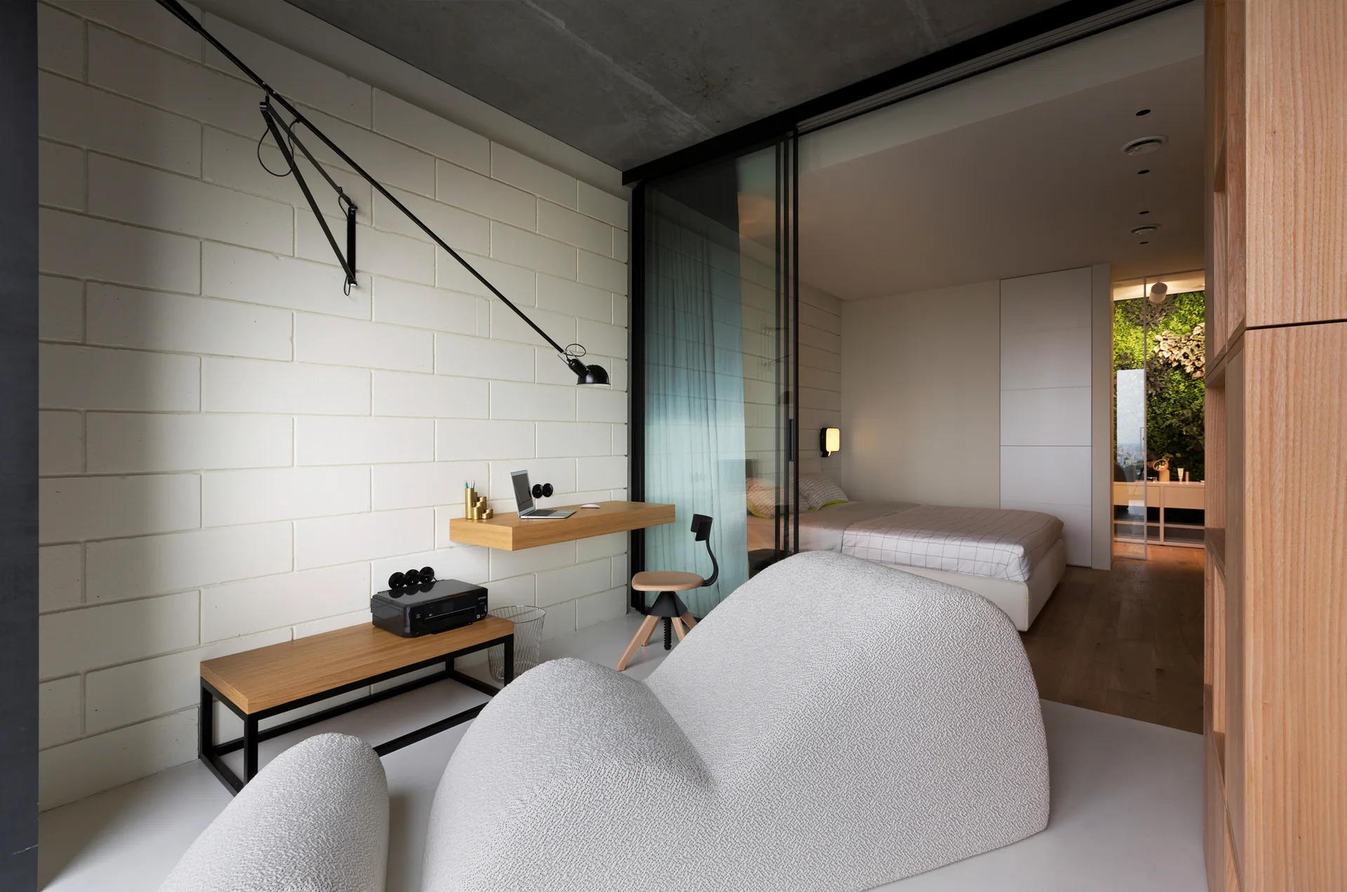 A modern bedroom office featuring a white painted brick wall with a floating desk, a sculptural lounge chair, and a sliding glass partition.