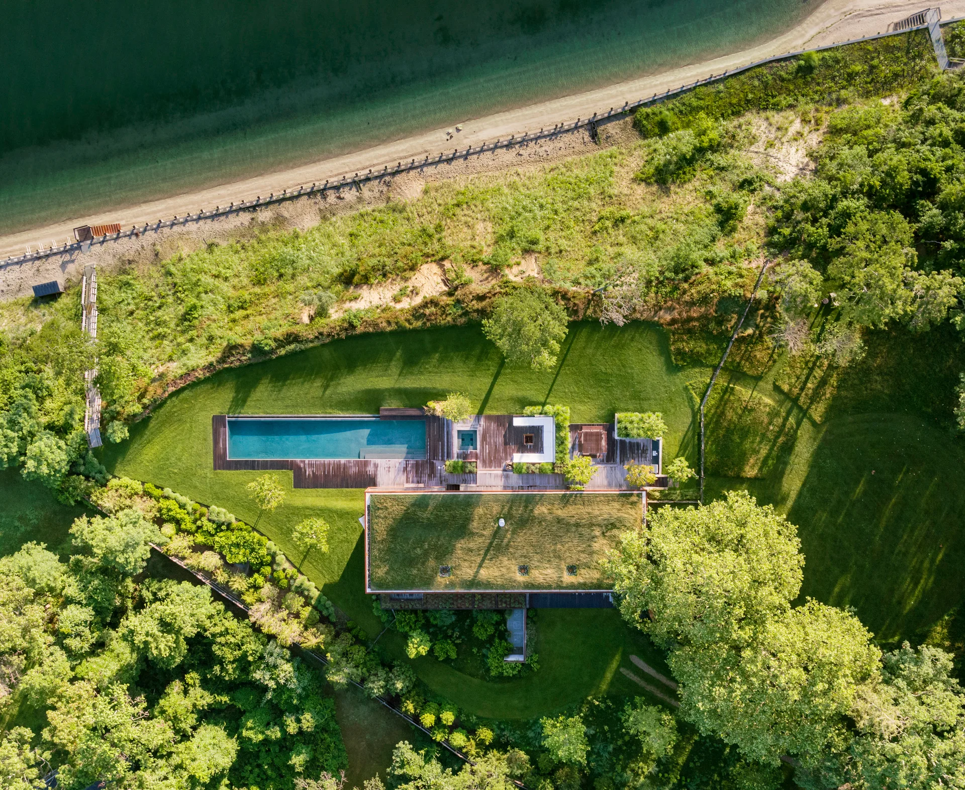 An aerial drone view of a modern coastal home featuring a green grass roof, a wooden deck, and a long blue swimming pool near a sandy beach.