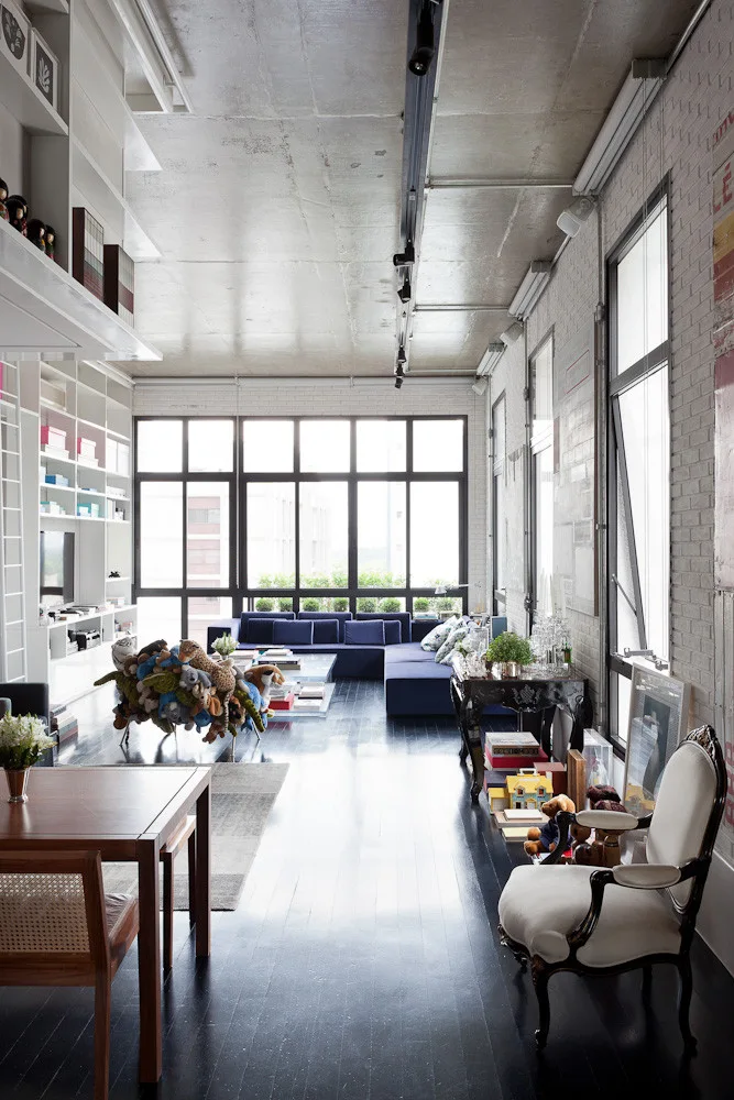 An industrial loft living room featuring an exposed concrete ceiling, painted white brick walls, a large blue sectional sofa, and dark wood floors.