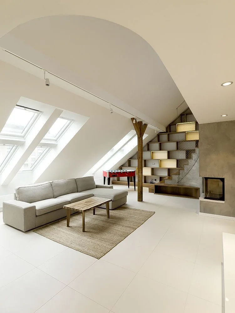 A bright loft living room featuring a grey sectional sofa, sloped skylights, and a wooden staircase with integrated open shelving.