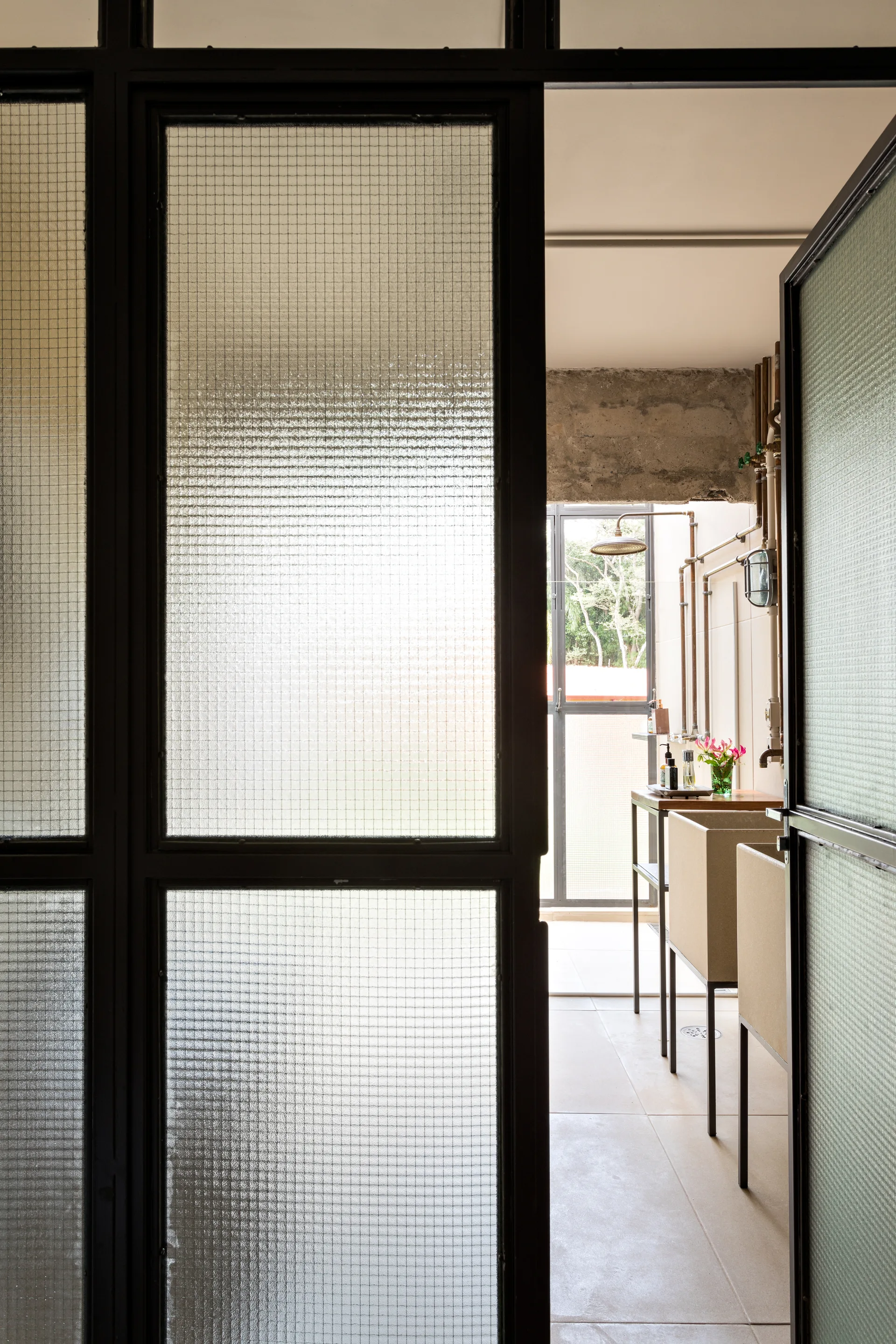An open black metal door with textured wire glass panels looks into a bathroom with exposed concrete, copper plumbing, and modern rectangular basins.