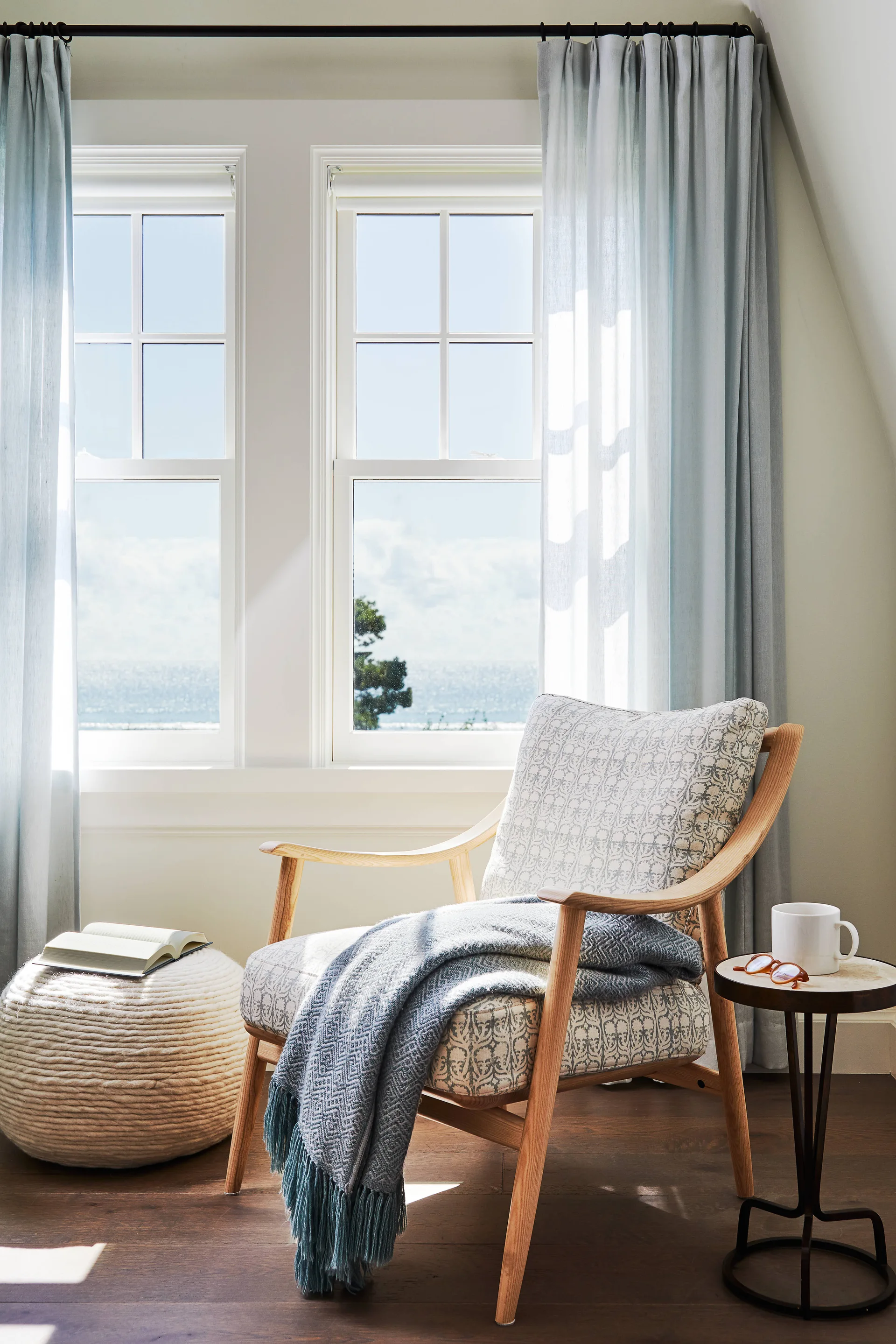 A patterned armchair sits in front of two double-hung windows with light blue curtains, alongside a textured pouf and small side table.