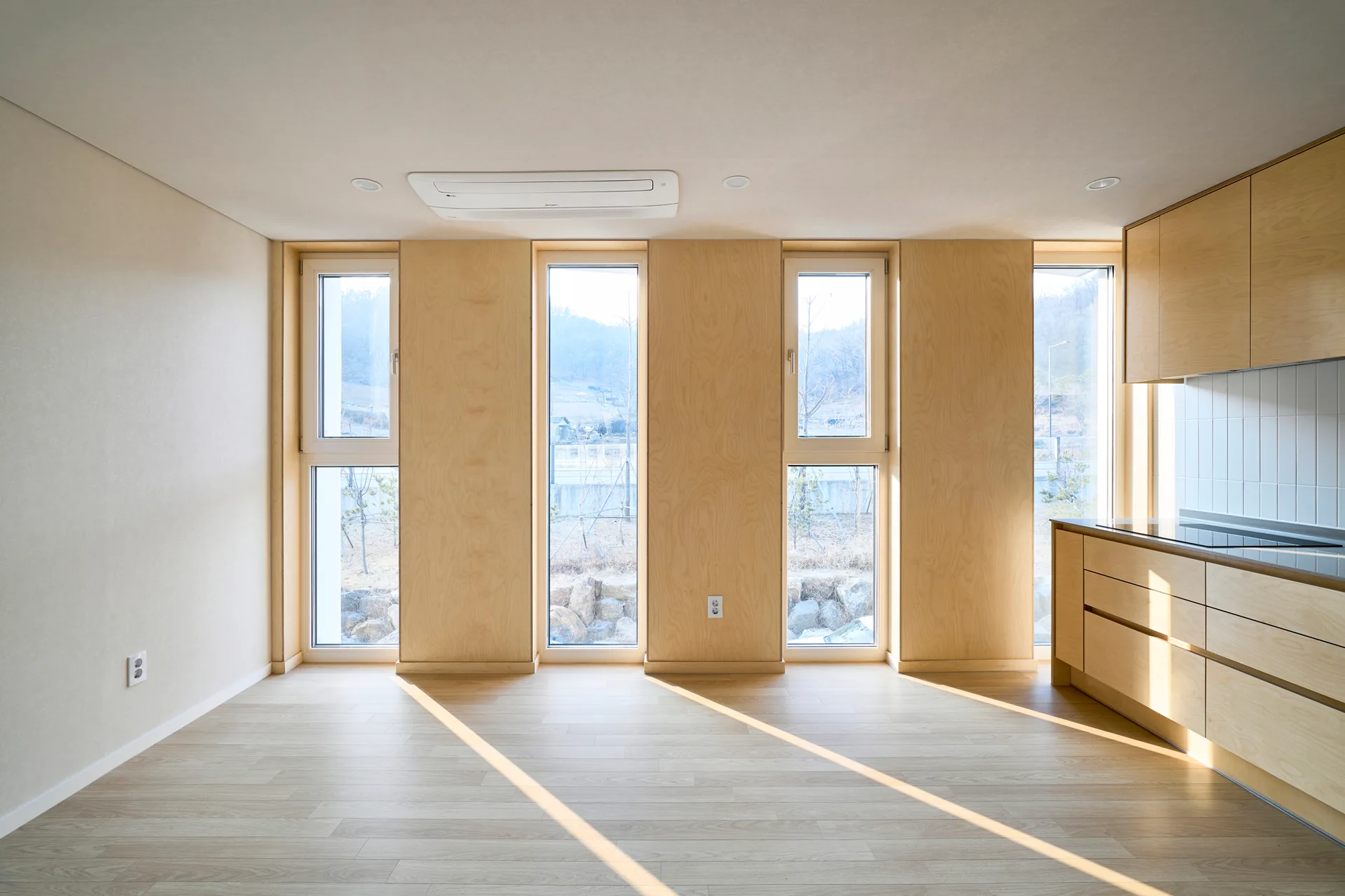 A minimalist kitchen features light wood cabinets, matching vertical wall panels between tall narrow windows, and sunlight streaming across wood floors.