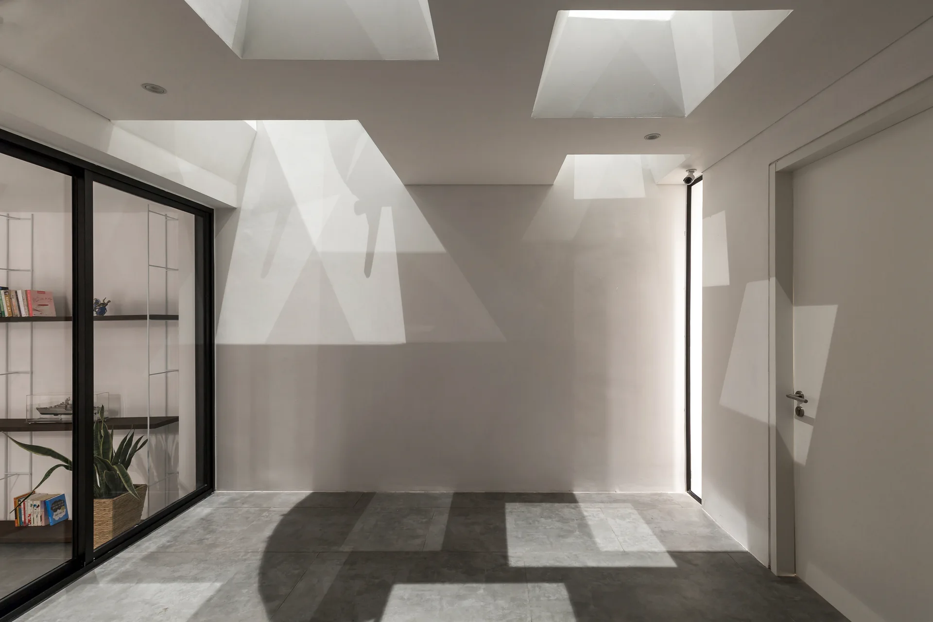 A minimalist white hallway features square ceiling skylights that cast dramatic geometric shadows across the walls and gray tile floor.