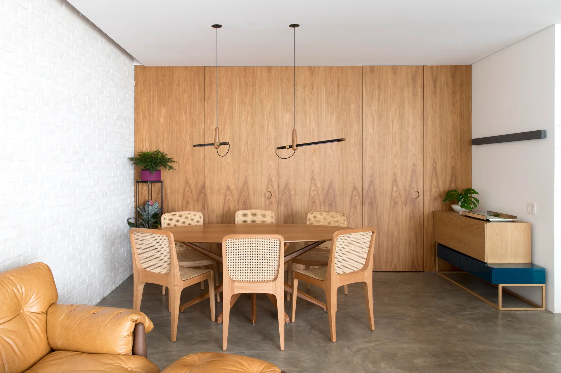 A modern dining room features an oval wood table and cane-back chairs positioned against a full-height wood paneled wall.