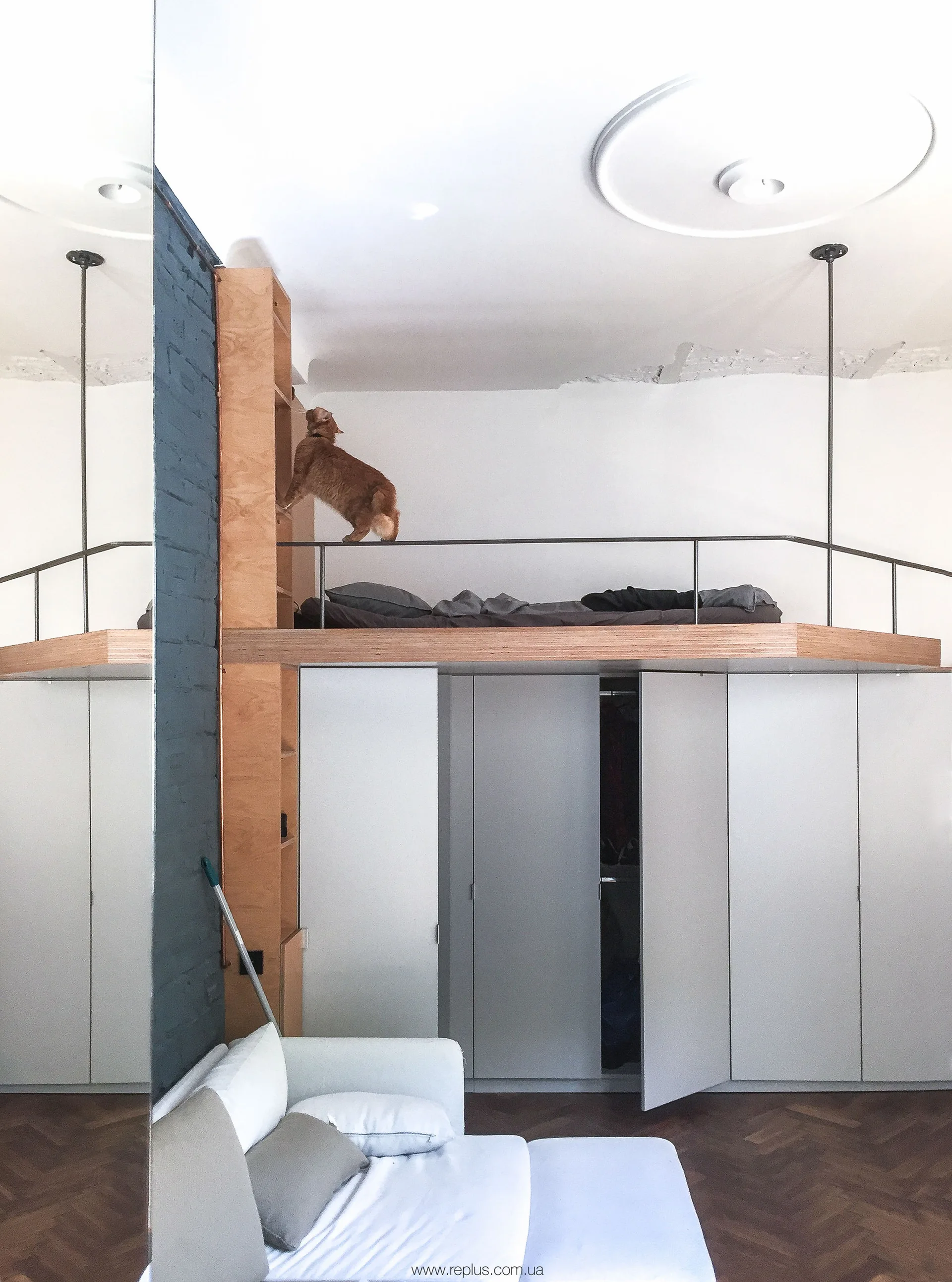 An elevated plywood loft bed with a thin metal railing built directly above a row of flush white wardrobes.