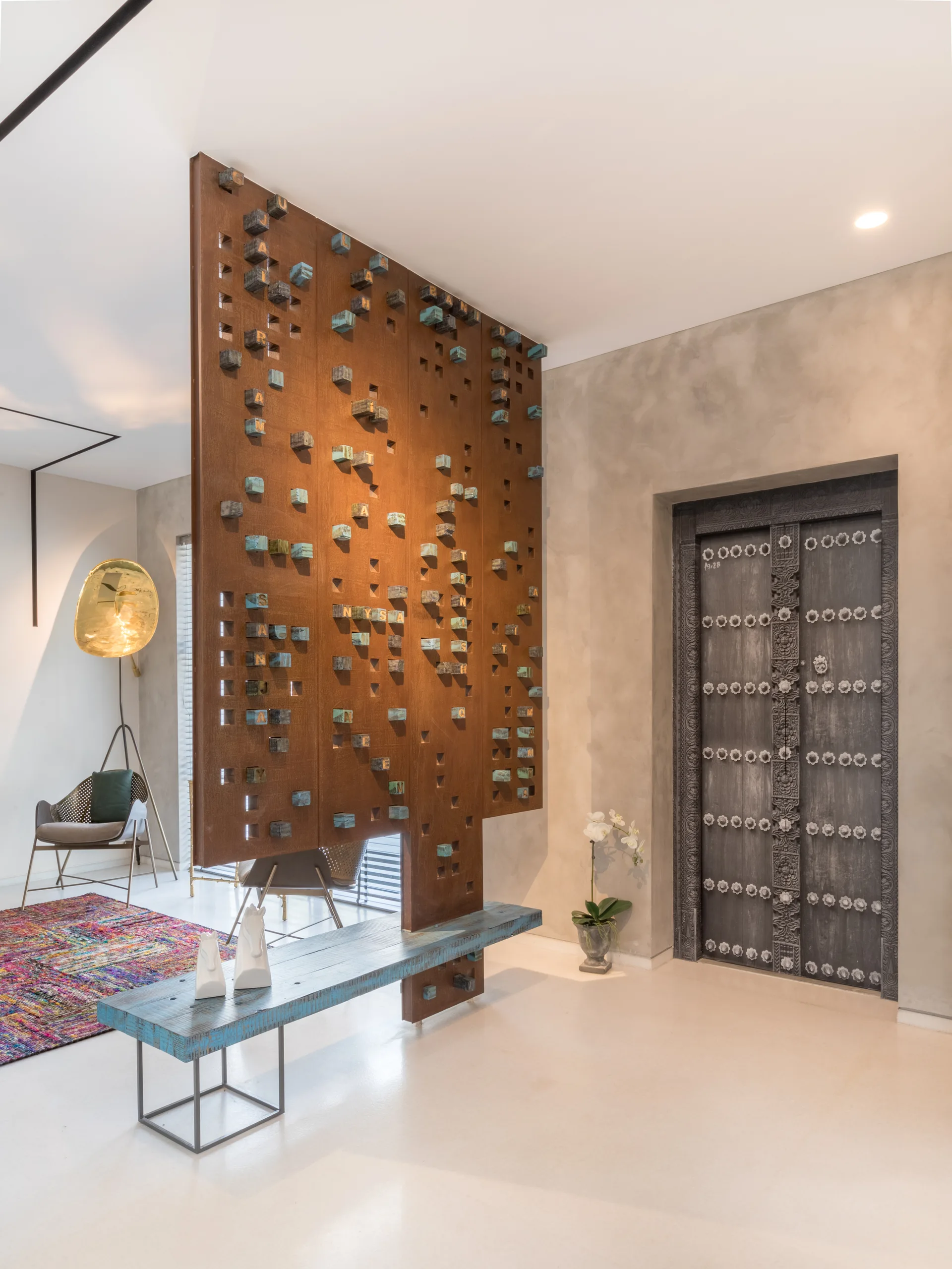 A modern entryway features a rusty metal room divider with geometric block details, a rustic blue wooden bench, and an ornate carved wooden door.